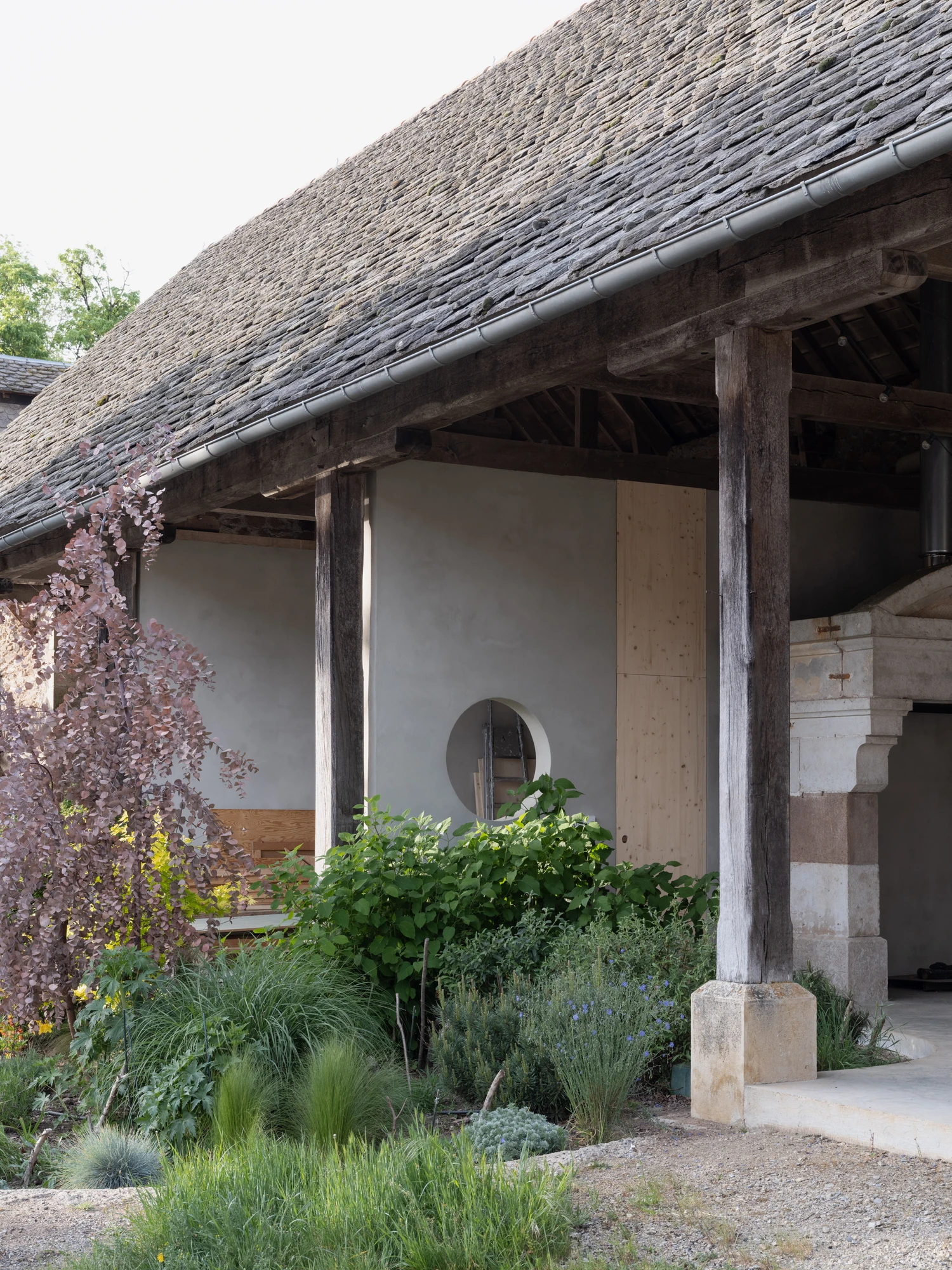 Farmhouse Renovation by Atelier AJO, photographed by Sandrine Iratçabal — The open barn from the courtyard garden, with a circular opening in the wall and the stone fireplace at right.