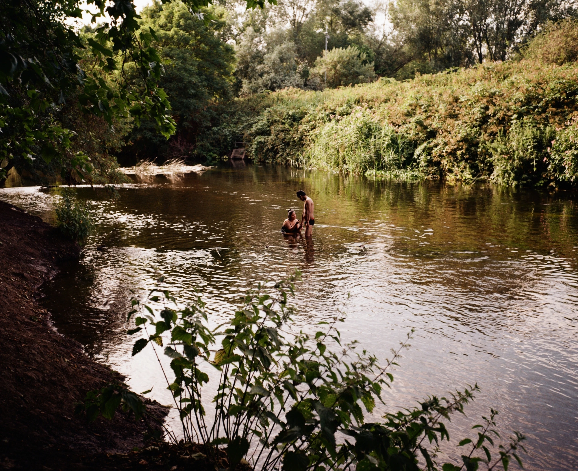 Along the River Lea by Mattia Corato, photographed by Mattia Corato — Hackney Marshes: two people standing in the river between overgrown banks.
