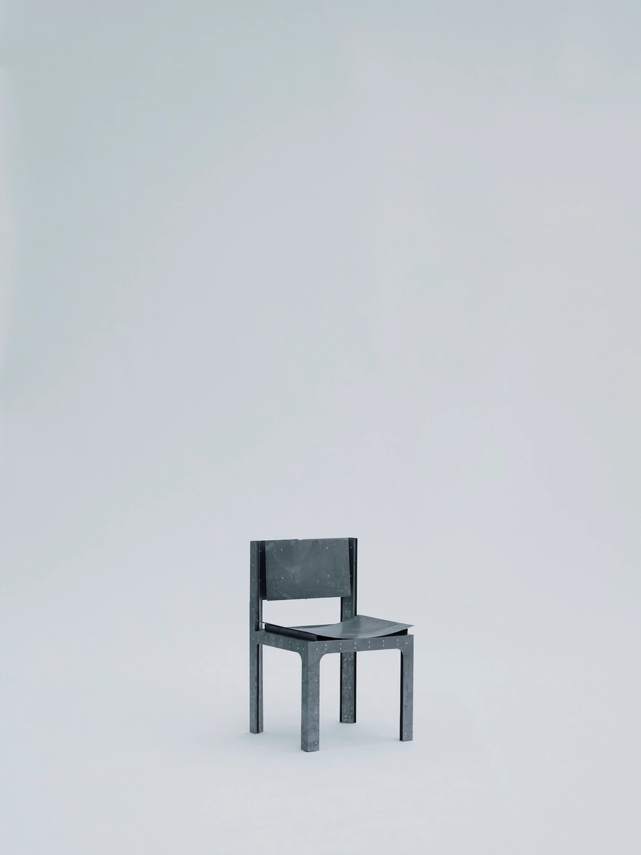 Trauer Tragen, Carrying Grief by Antonia Fischer, photographed by Maya Matsuura, Antonia Fischer — A vertical crop of the studio portrait, the riveted steel chair standing small against a grey backdrop.