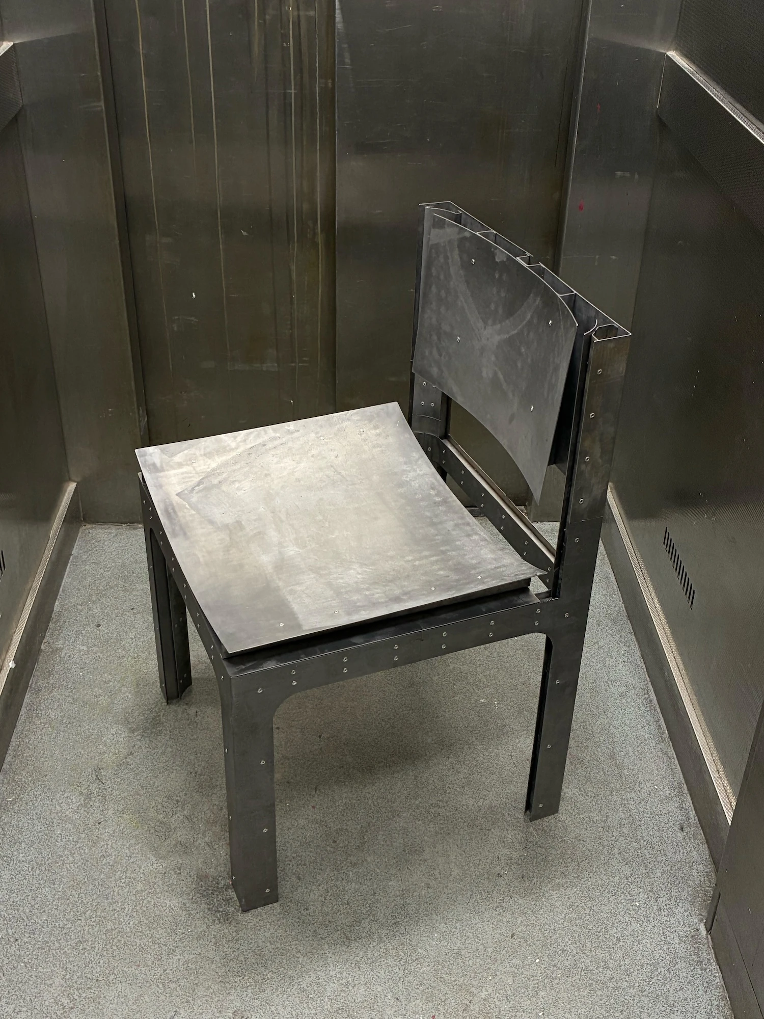 Trauer Tragen, Carrying Grief by Antonia Fischer, photographed by Maya Matsuura, Antonia Fischer — The chair photographed in colour inside a steel-lined lift or booth, rivets running along the frame.
