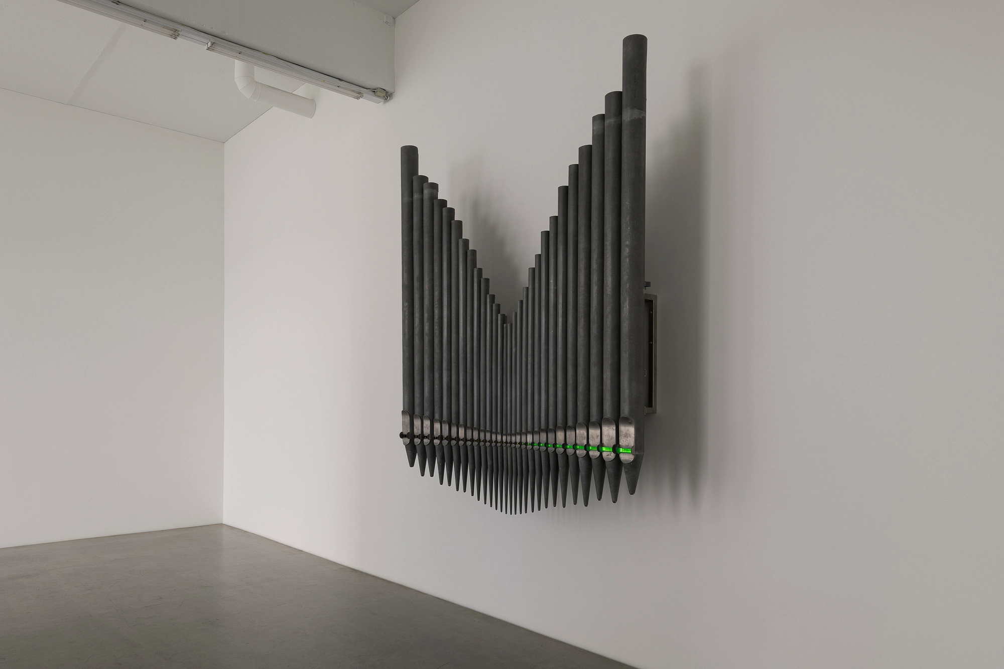 Half-Life of Grace by Jeremy Shaw, photographed by Stefan Korte — An angled view of the organ-pipe sculpture, its rows receding along the wall with the green glow catching each mouth.