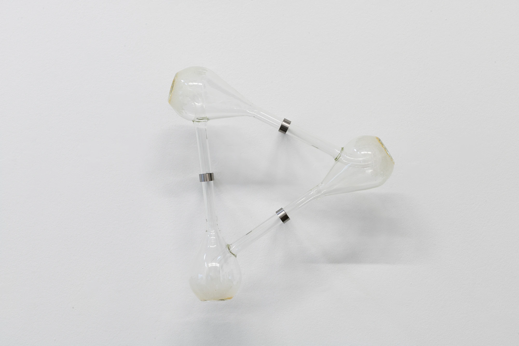 Half-Life of Grace by Jeremy Shaw, photographed by Stefan Korte — The Distance Between Infinite Folds is Still You in full, three glass bulbs linked by tubes and stainless steel collars into a triangle.