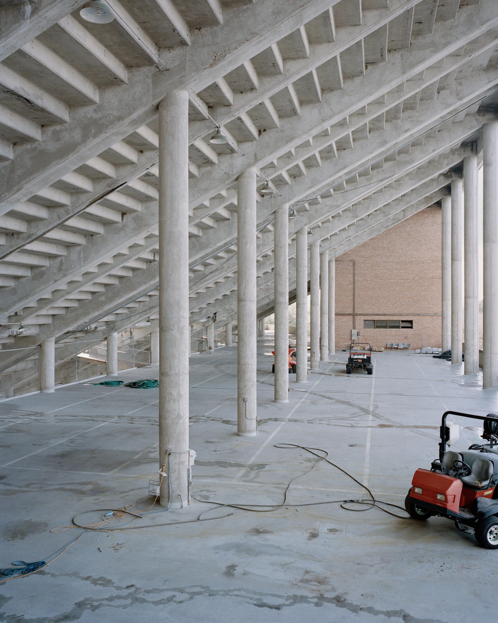 Houston, Untitled by Rory Gardiner, photographed by Rory Gardiner — 29.716585, -95.410160: beneath a stadium's stands, rows of concrete pillars repeat, with small maintenance vehicles and cables left on the floor.