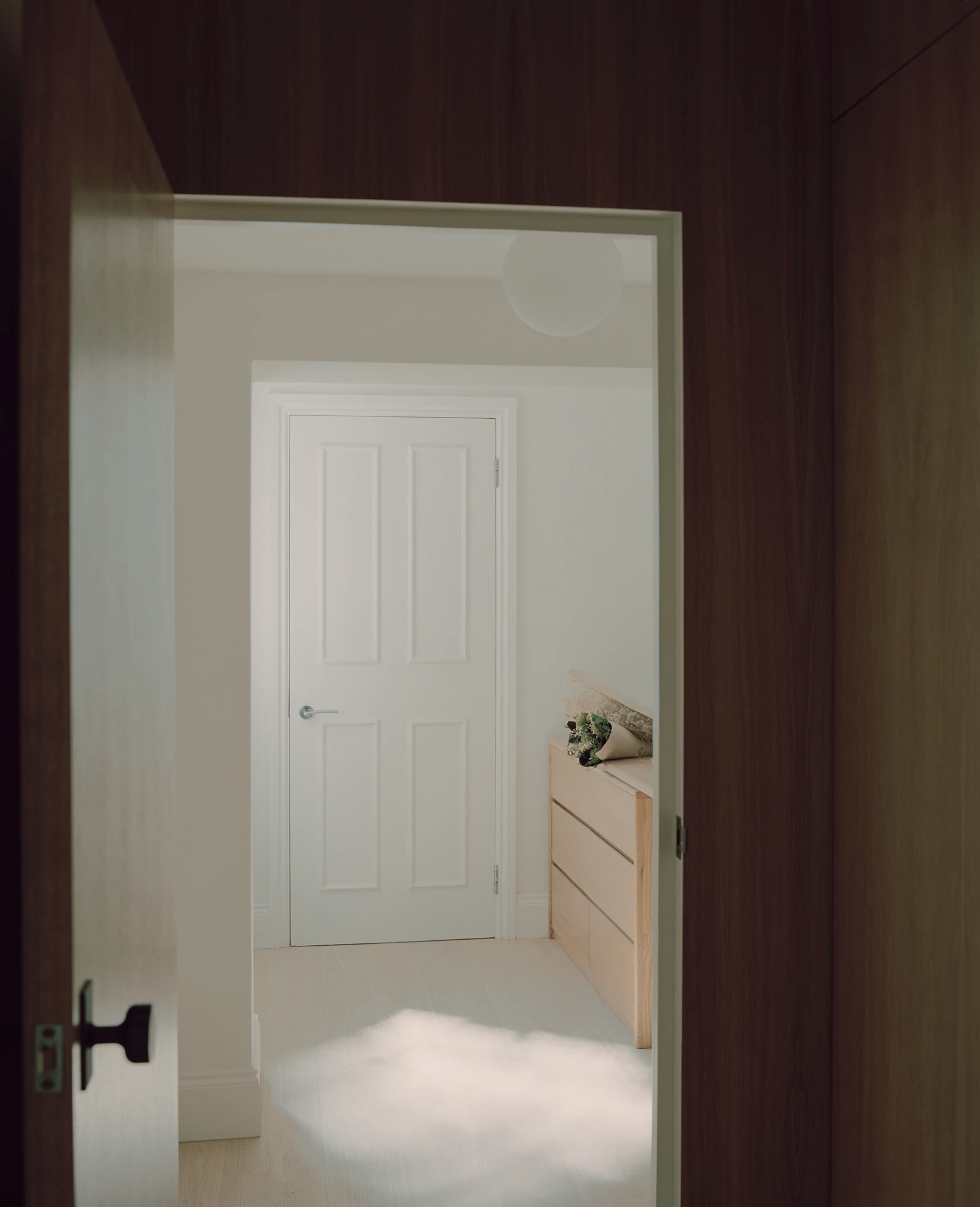 Hampstead House by Twelve Forty One, photographed by Willem Pab — A view across a landing framed in dark wood toward a white panelled door, a pale oak chest of drawers lit by the window.