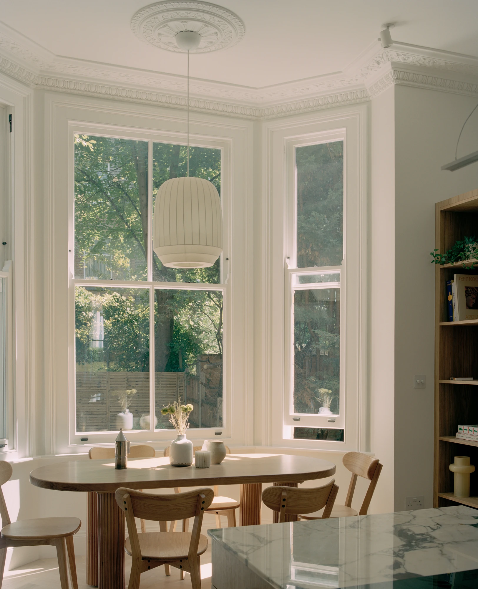Hampstead House by Twelve Forty One, photographed by Willem Pab — The dining table set into a bay window beneath a moulded ceiling rose, an oak bookcase standing to one side.