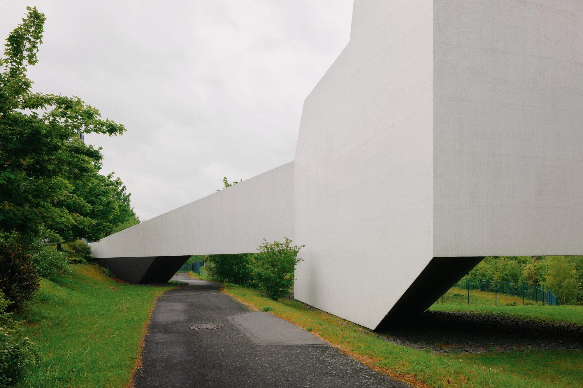 Autobahnkirche Siegerland by schneider+schumacher, photographed by David Altrath — Exterior view where a paved path passes beneath the raised white access bridge between canted walls.