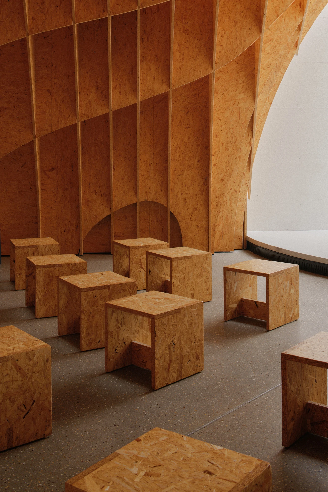 Autobahnkirche Siegerland by schneider+schumacher, photographed by David Altrath — Interior view of cube-shaped wooden stools on the floor before the ribbed timber wall and a low arched opening.