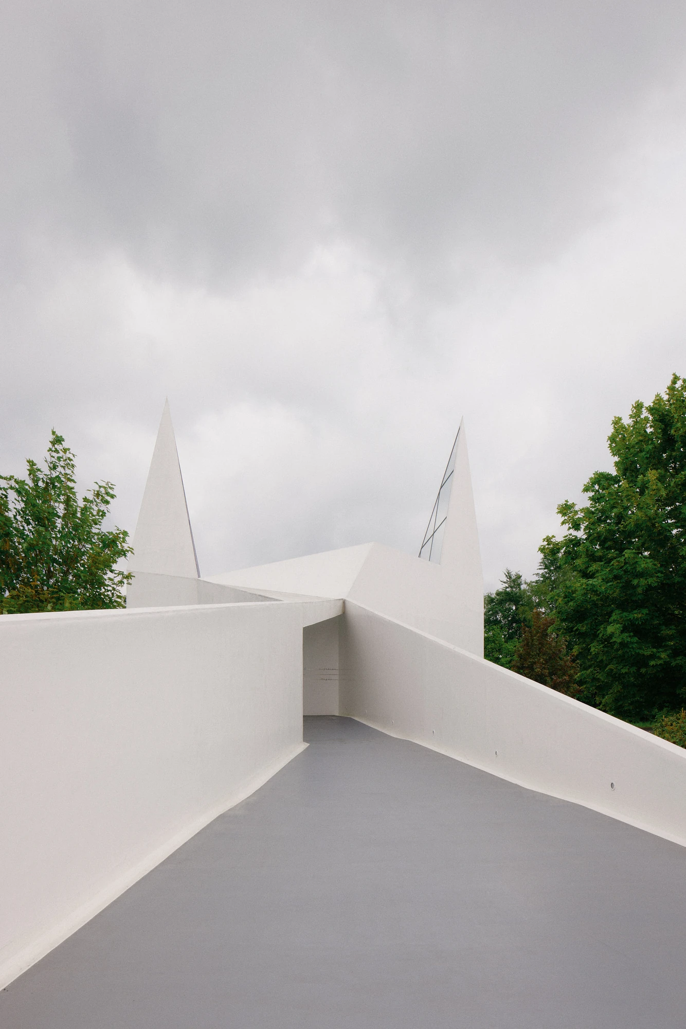 Autobahnkirche Siegerland by schneider+schumacher, photographed by David Altrath — Exterior view along the grey rooftop walkway between white parapets leading toward the two pointed spires.