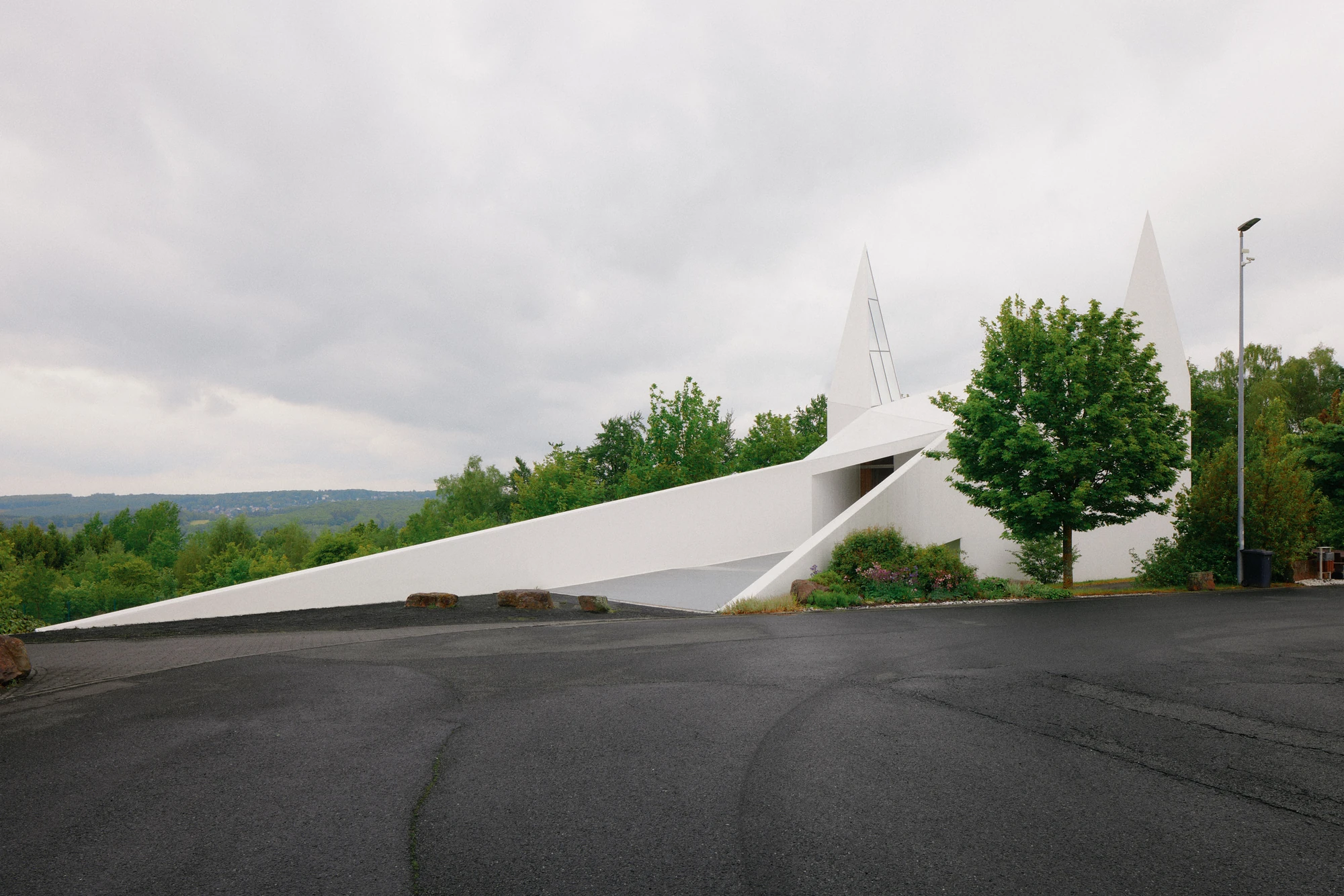 Autobahnkirche Siegerland by schneider+schumacher, photographed by David Altrath — Exterior view of the long white volume and its sloping access ramp stretching across the tarmac toward the two spires.