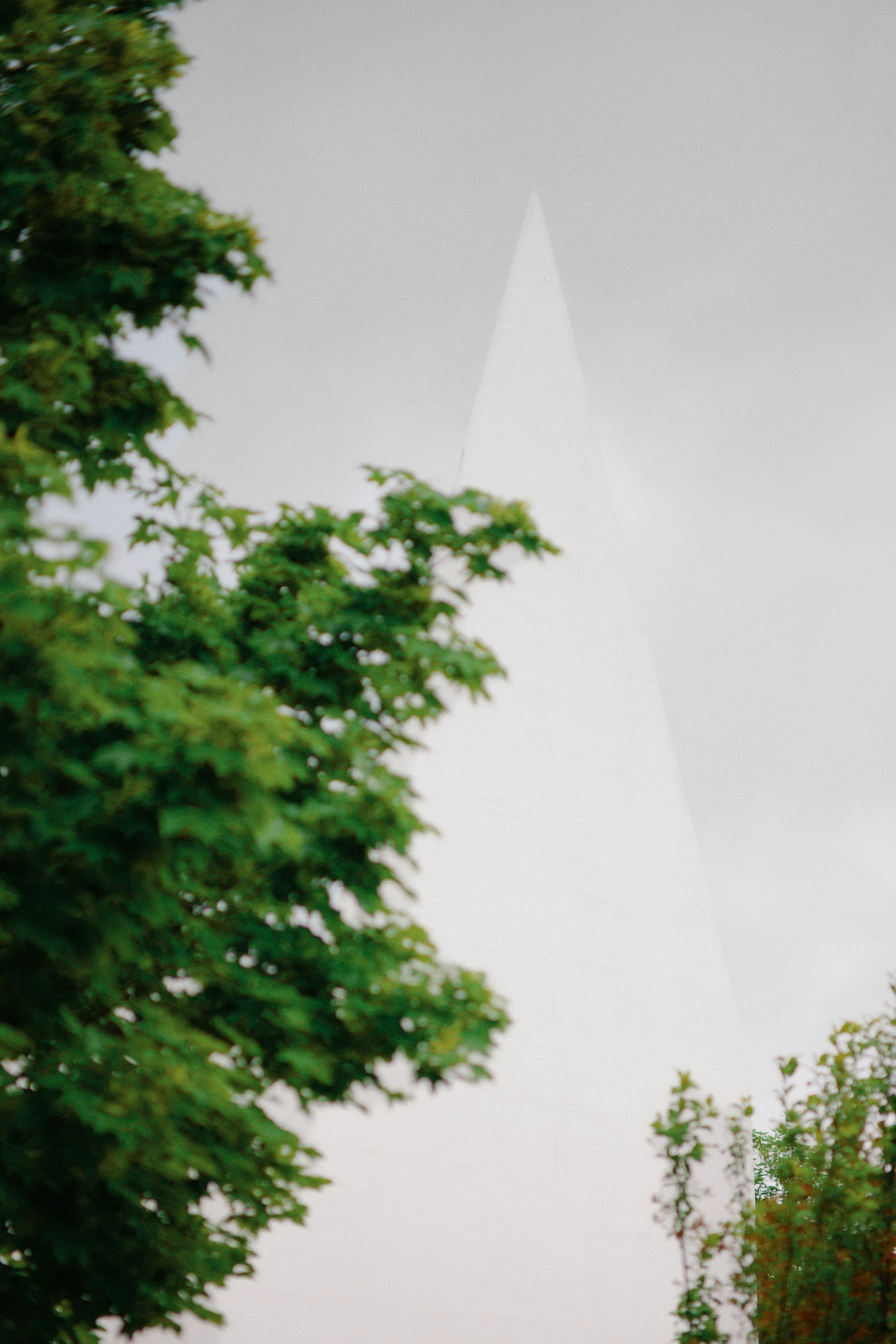 Autobahnkirche Siegerland by schneider+schumacher, photographed by David Altrath — Exterior view of a single white spire seen past green maple foliage against a grey sky.