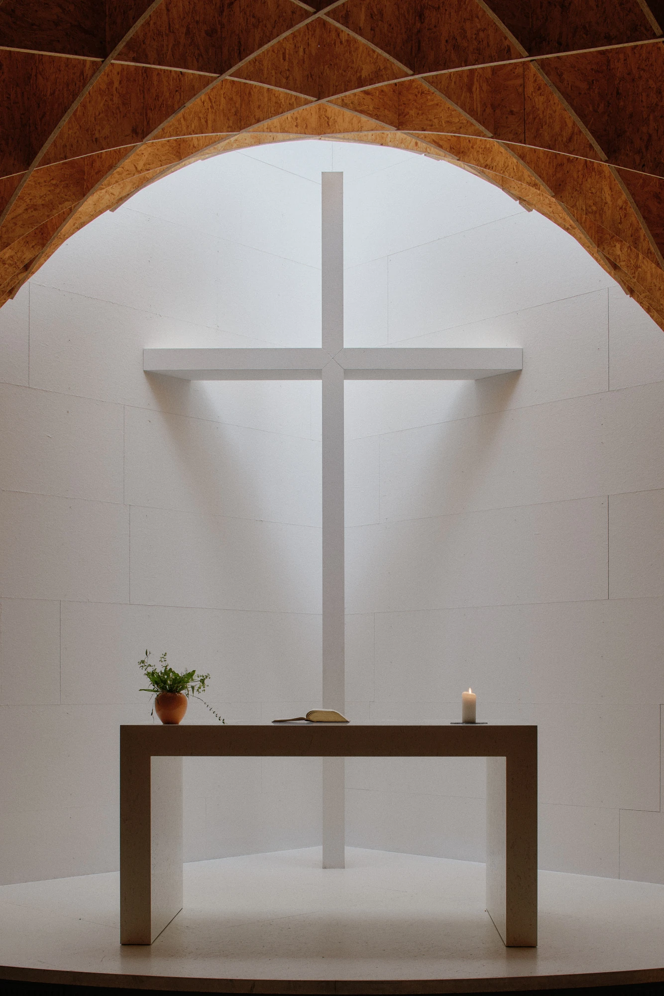 Autobahnkirche Siegerland by schneider+schumacher, photographed by David Altrath — Interior view of the white altar table with a candle and a potted plant below the cross on the apse wall.