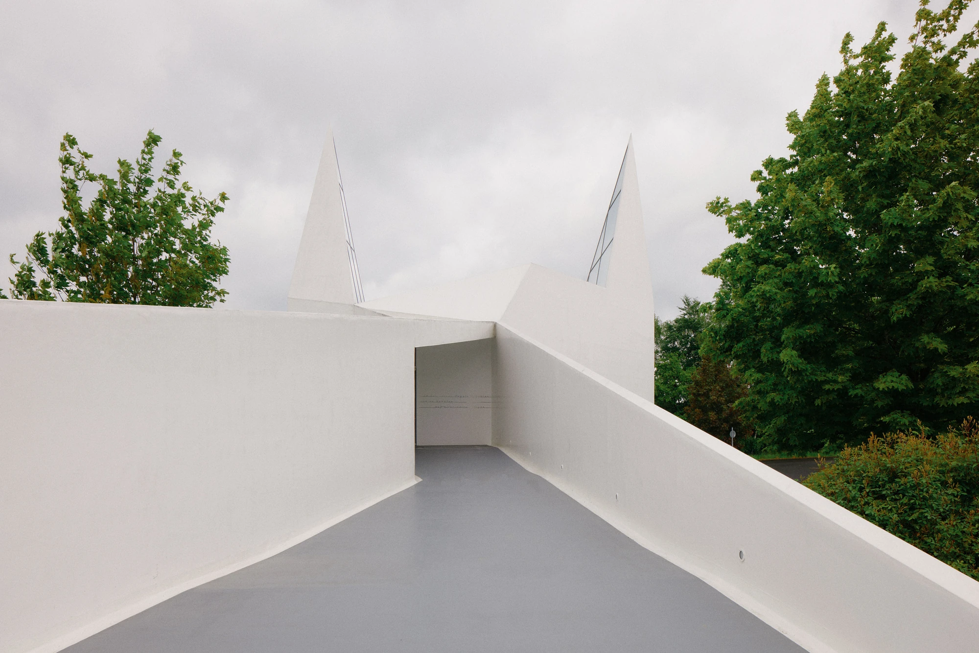 Autobahnkirche Siegerland by schneider+schumacher, photographed by David Altrath — Exterior view of a raised white walkway leading toward the church between angular parapet walls.