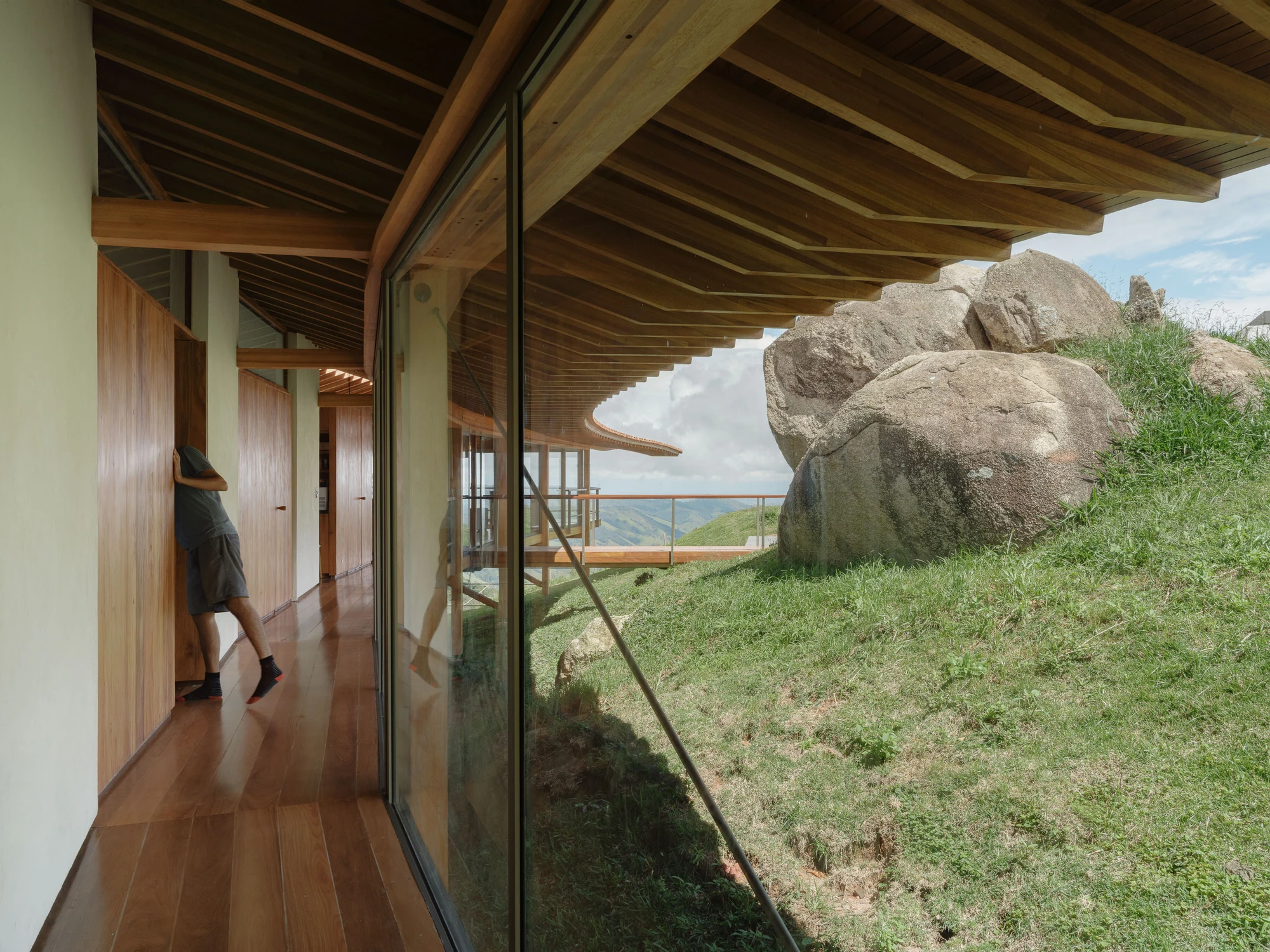 Casa Fazenda Bocaina by Arquipélago Arquitetos + Mariana Caires, photographed by Pedro Kok — Interior glazed corridor with a person at left and boulders close against the glass outside.