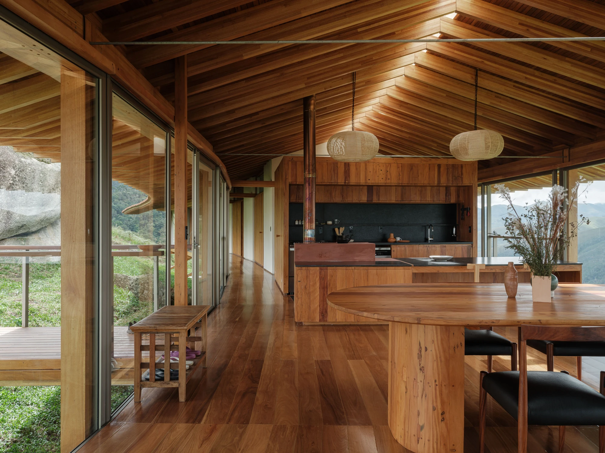 Casa Fazenda Bocaina by Arquipélago Arquitetos + Mariana Caires, photographed by Pedro Kok — The kitchen and dining area with pendant lamps, a stove flue and the glazed wall onto the hills.