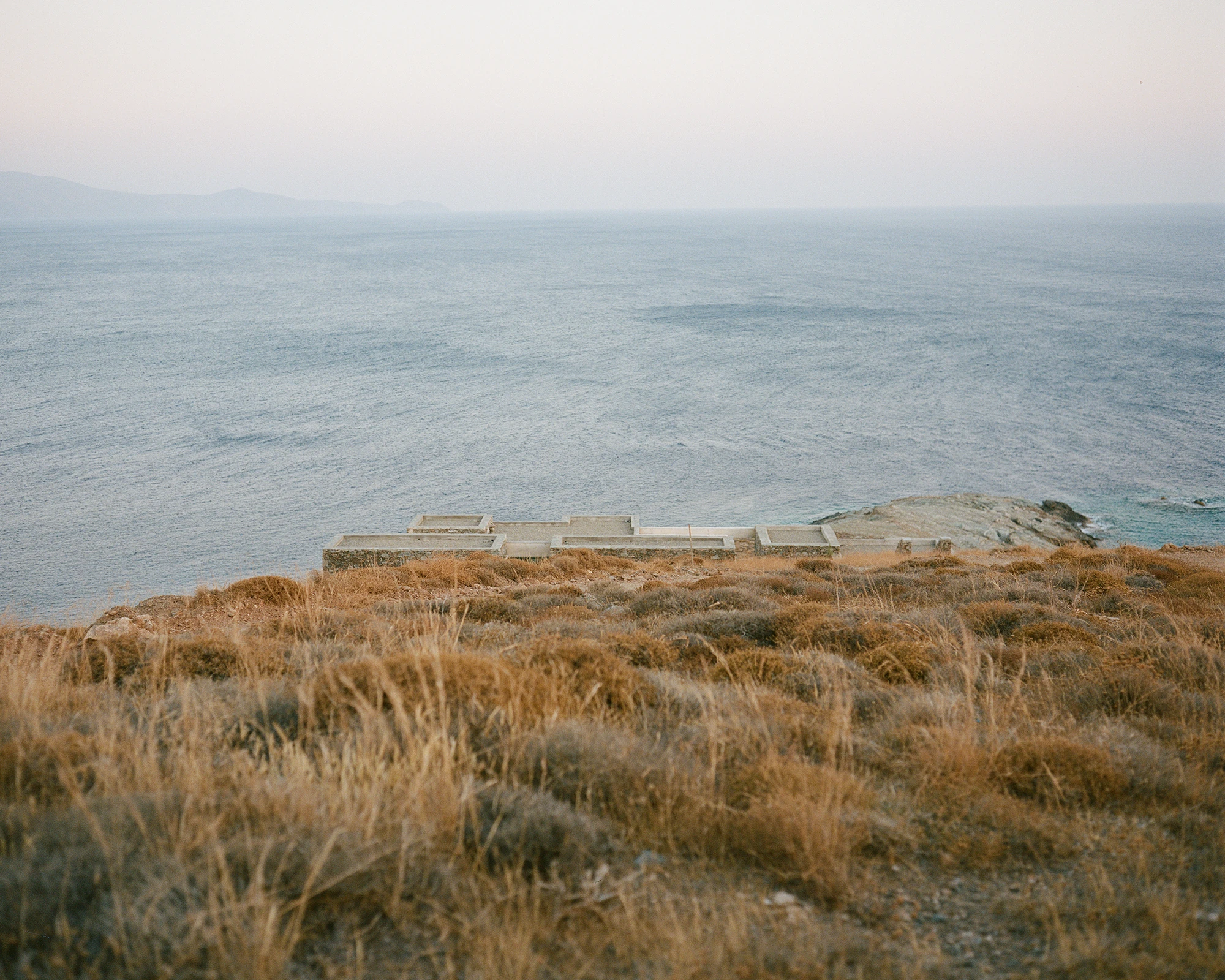 House Thermia by RAUM, photographed by Benoit Chailleux — The house across the hillside, low above the dry grass at Thermia.