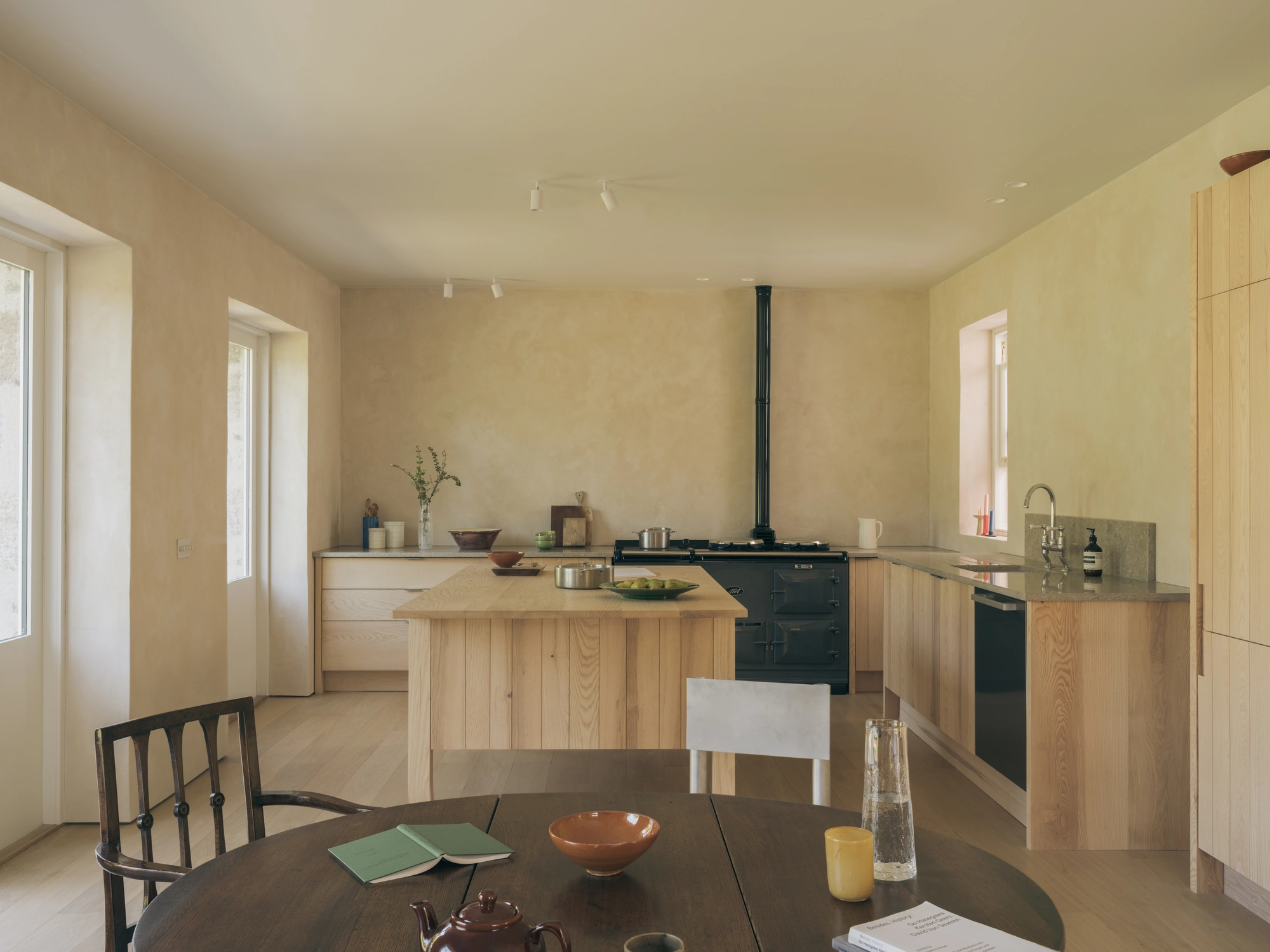 Cairngorm Cottage by Architecture Office, photographed by Richard Gaston — The full kitchen seen from the dining table, ash cabinetry and stone worktops under clay-plaster walls.