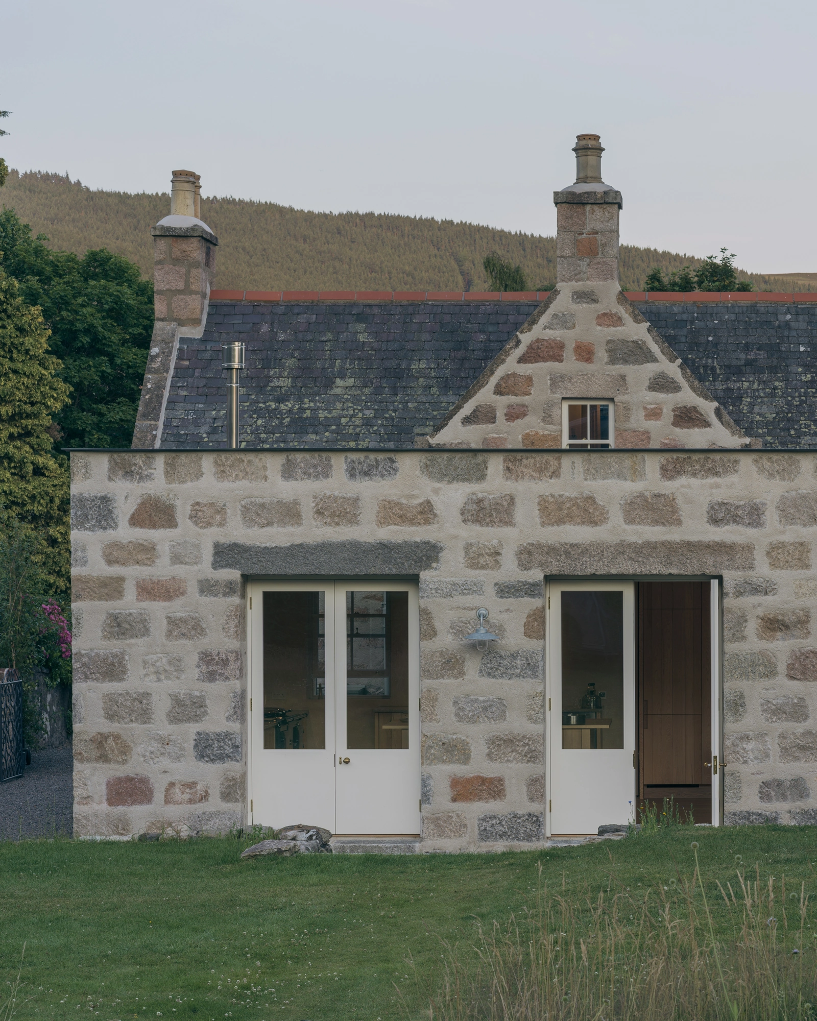 Cairngorm Cottage by Architecture Office, photographed by Richard Gaston — A closer view of the granite extension, two glazed doors in the thick wall below the original cottage and its chimneys.