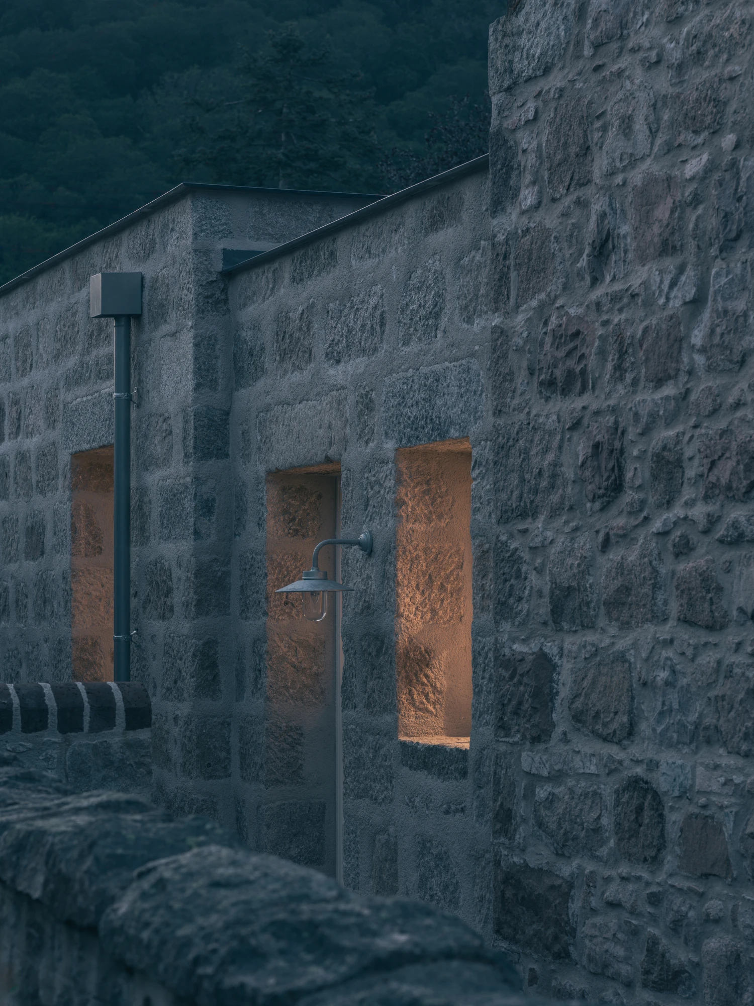 Cairngorm Cottage by Architecture Office, photographed by Richard Gaston — The side wall at dusk, deep-set openings lit in the granite with a rainwater downpipe running between them.