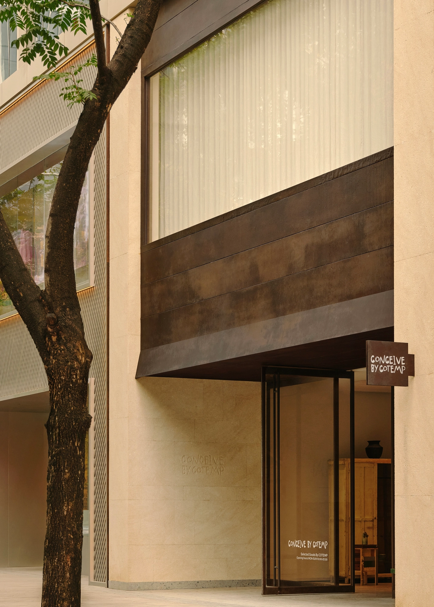 Conceived by Cotemp by ONOAA STUDIO, photographed by Jonathan Leijonhufvud — The full street front, the rusted iron canopy folding out above the entrance and the shop name on the pale mineral-rendered wall beside a tree.