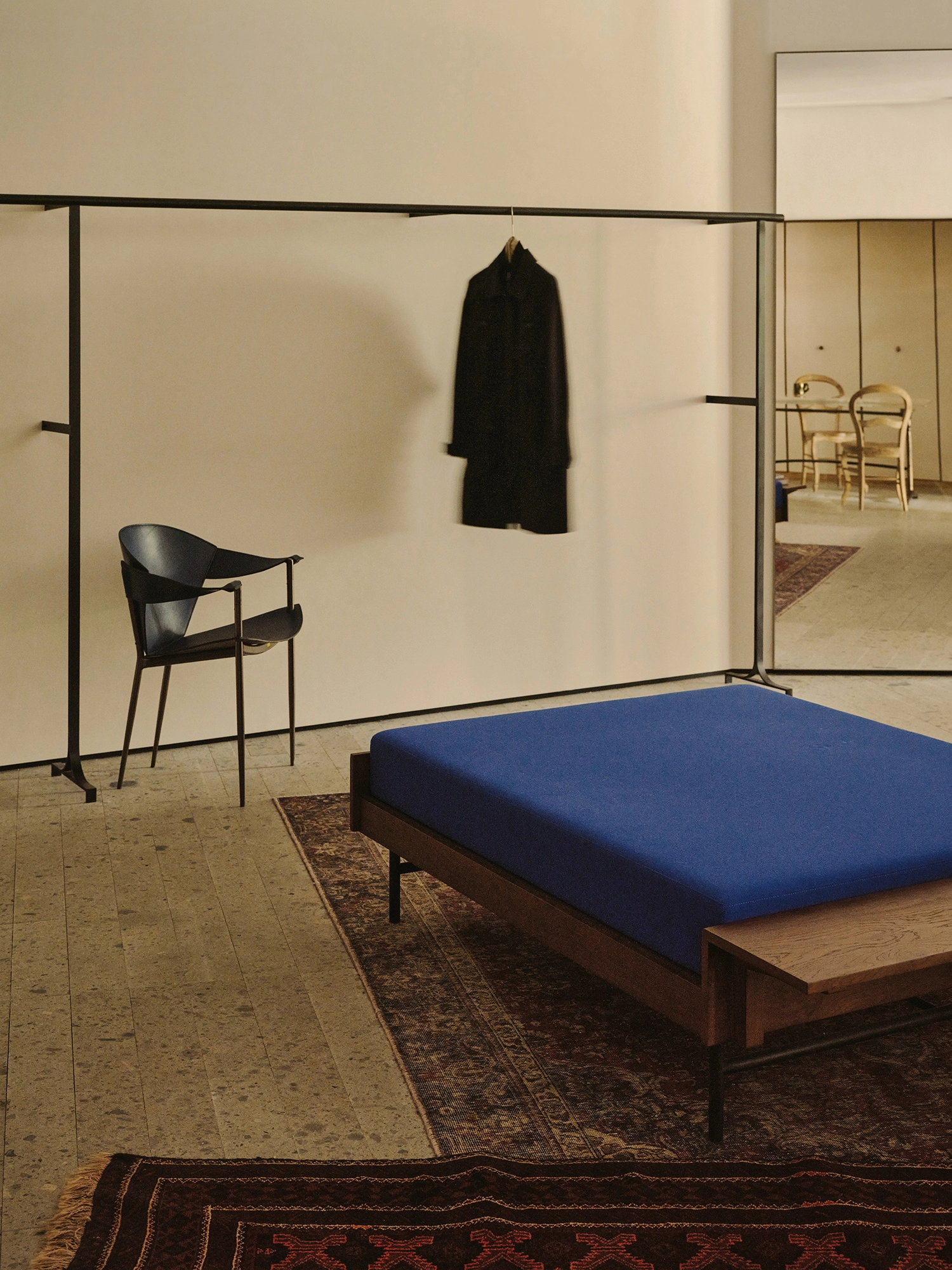 Conceived by Cotemp by ONOAA STUDIO, photographed by Jonathan Leijonhufvud — A coat on a wall rail above a daybed and a chair, with a mirror reflecting the room beyond.