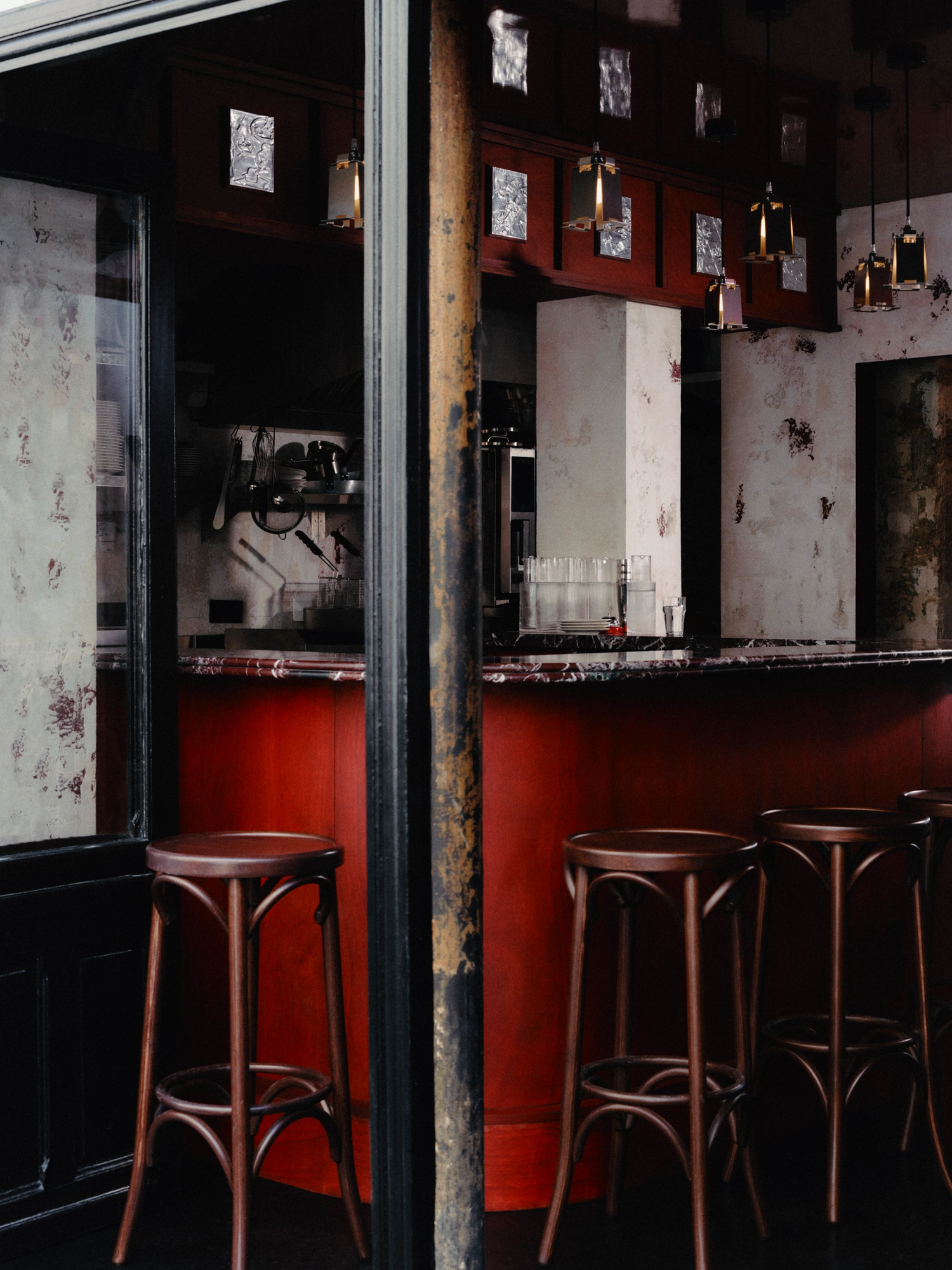 WINONA by Romain Basile Petrot, photographed by Romain Roucoules, Carla Beaujardin — The counter seen past a column, its deep red marble top over the wooden base, custom lights by Petrot strung above the stools.