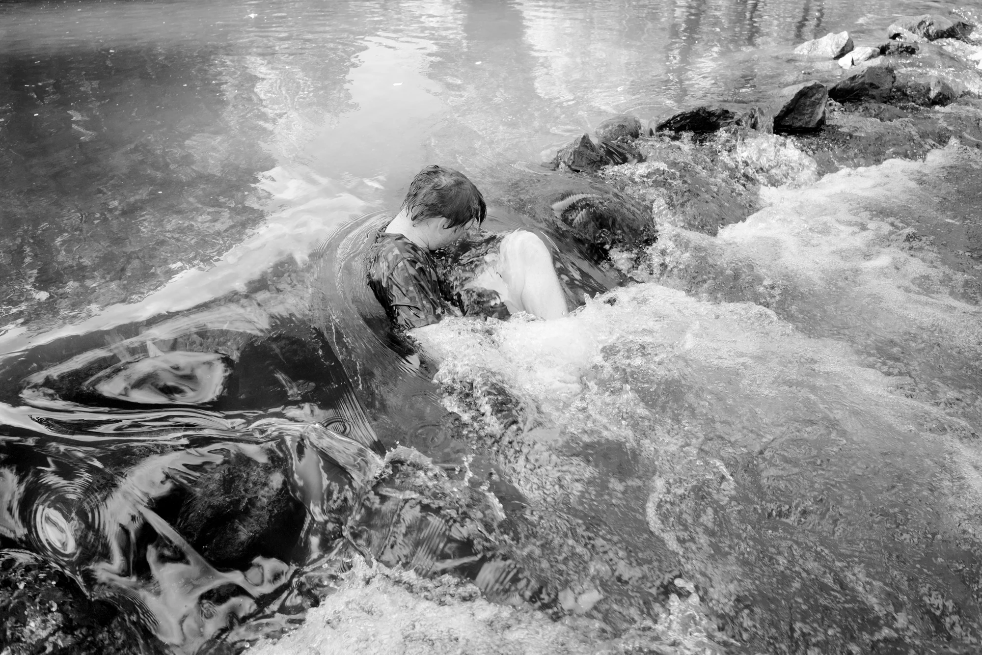 Down by the Hudson by Caleb Stein, photographed by Caleb Stein — Shamus in Embryo Pose (2022), a figure curled tight in the rushing white water above a low weir