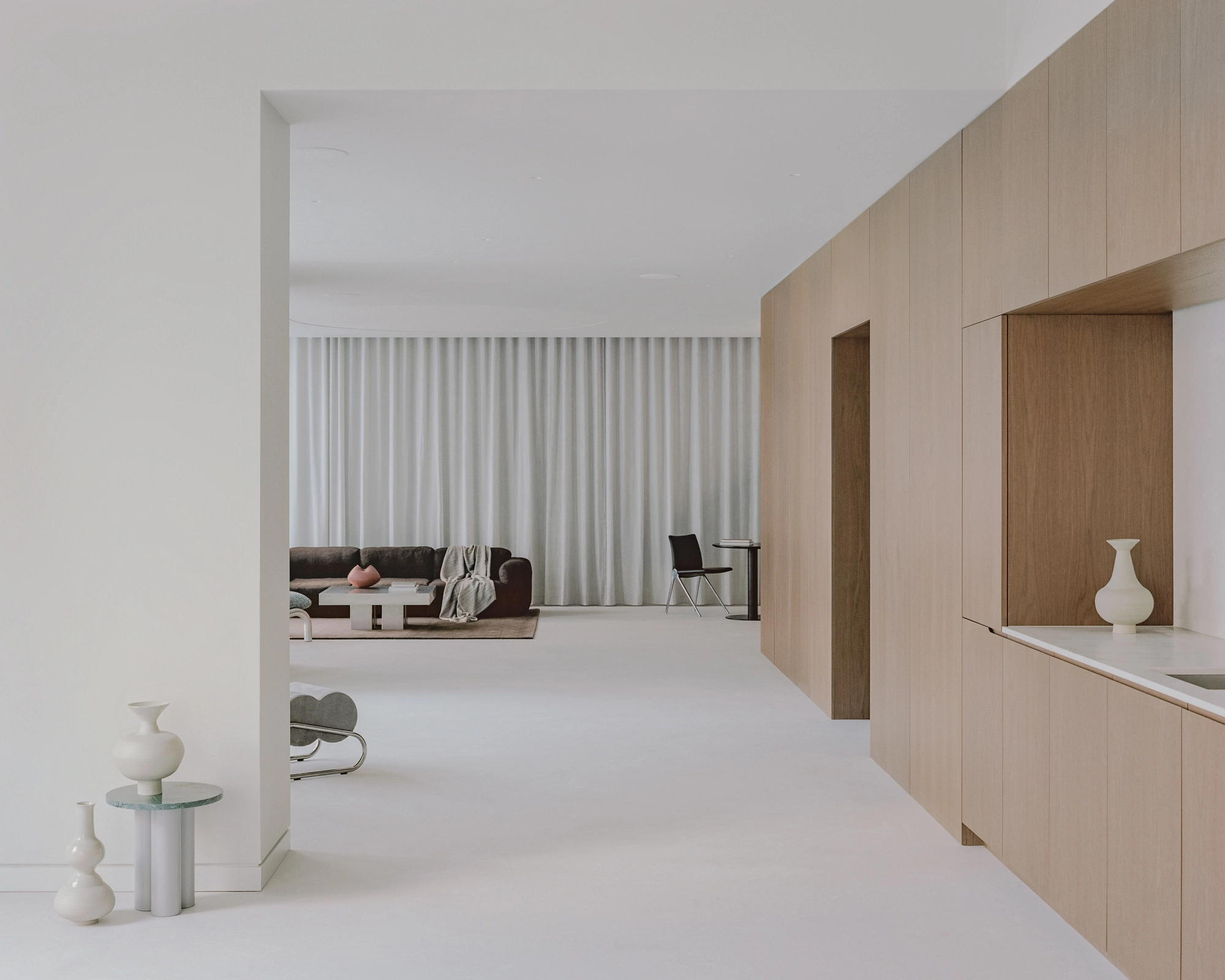Oval House by Actual Office, photographed by Nick Dearden — Looking past a pale wall of cupboards toward the living area, with ceramic vessels on a side table in the foreground.