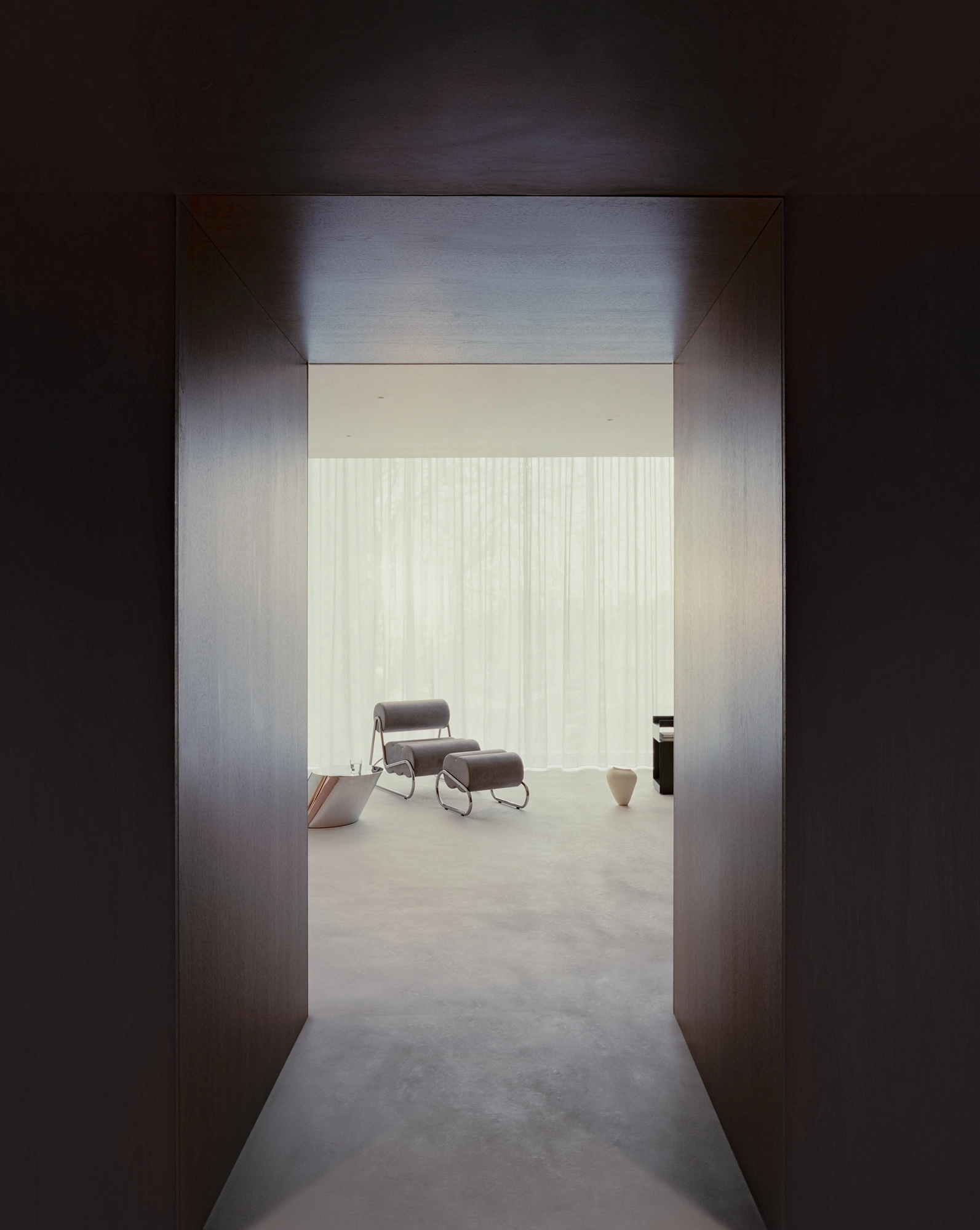 Oval House by Actual Office, photographed by Nick Dearden — View through a deep walnut threshold into the bright main space, with a grey chair and polished chrome side table by the sheer curtains.