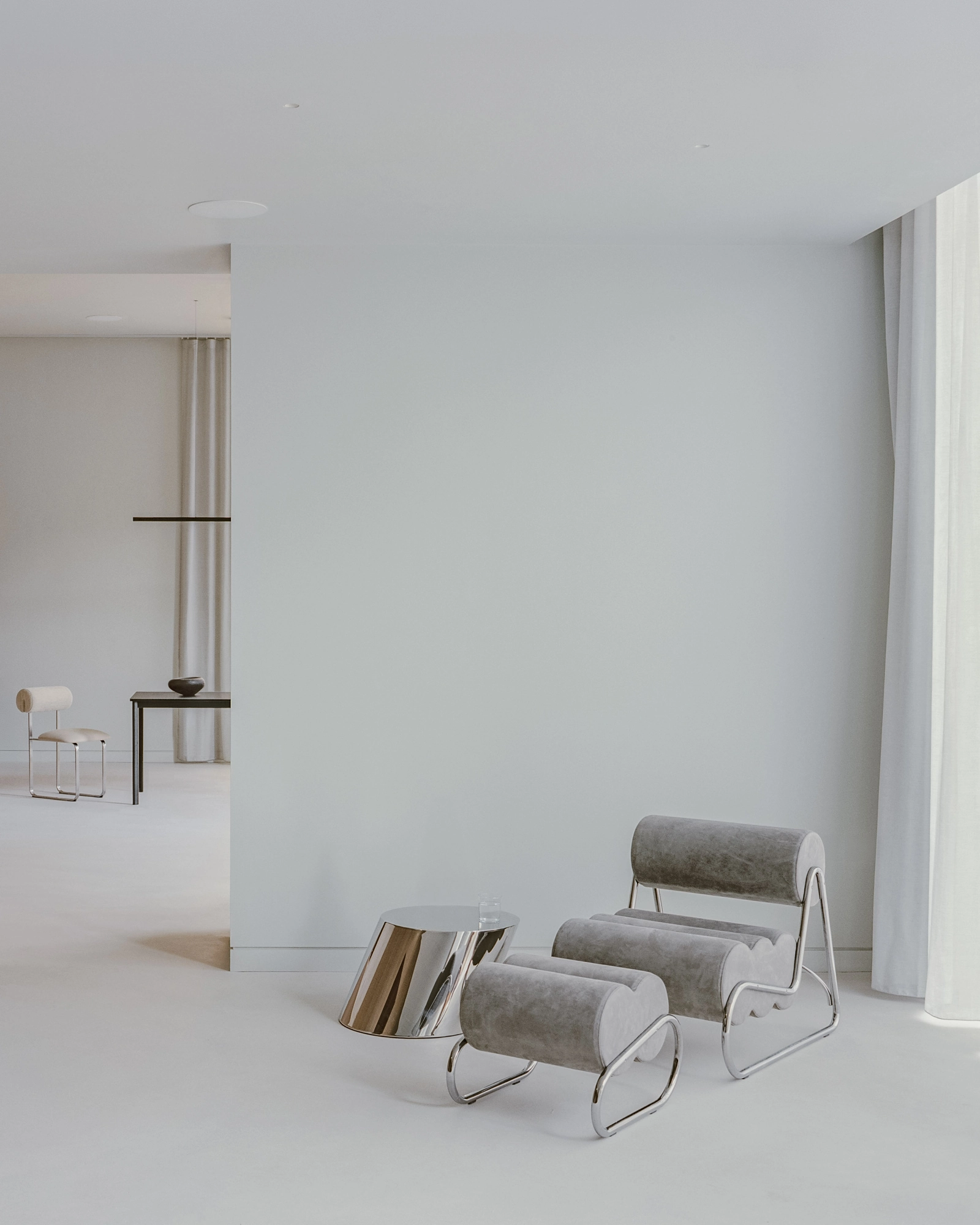 Oval House by Actual Office, photographed by Nick Dearden — A grey tubular lounge chair beside a polished chrome drum table, set against a pale wall with sheer curtains to the right.
