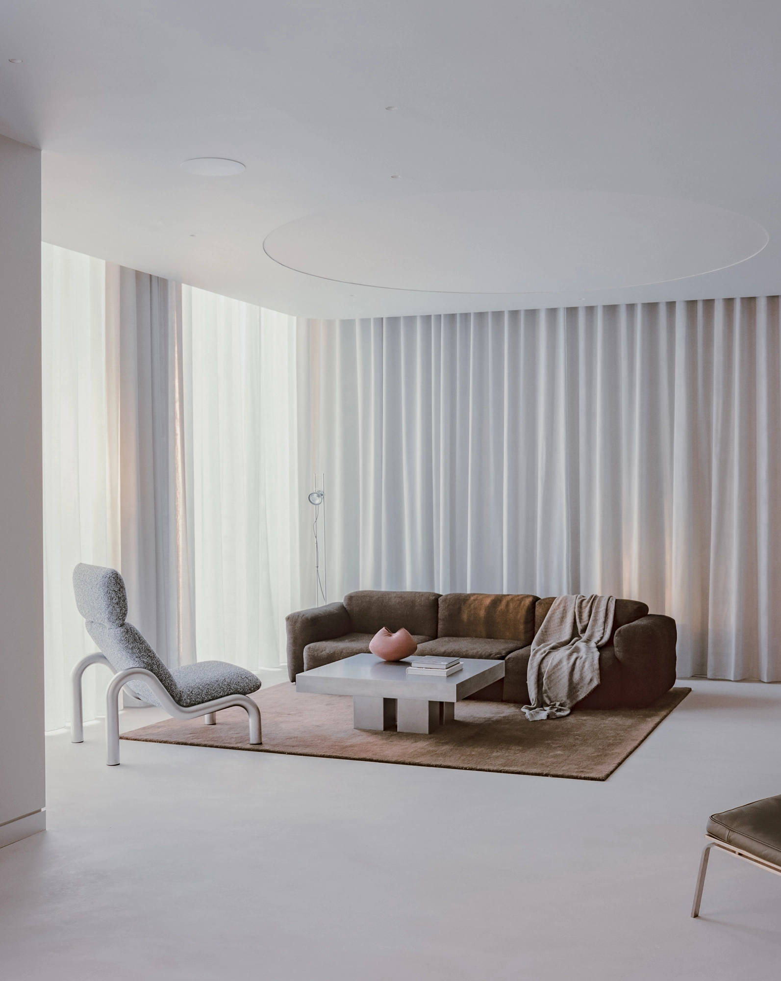 Oval House by Actual Office, photographed by Nick Dearden — The living area with a pale chaise, dark brown sofa and low square table on a rug, sheer curtains behind and an oval recess above.