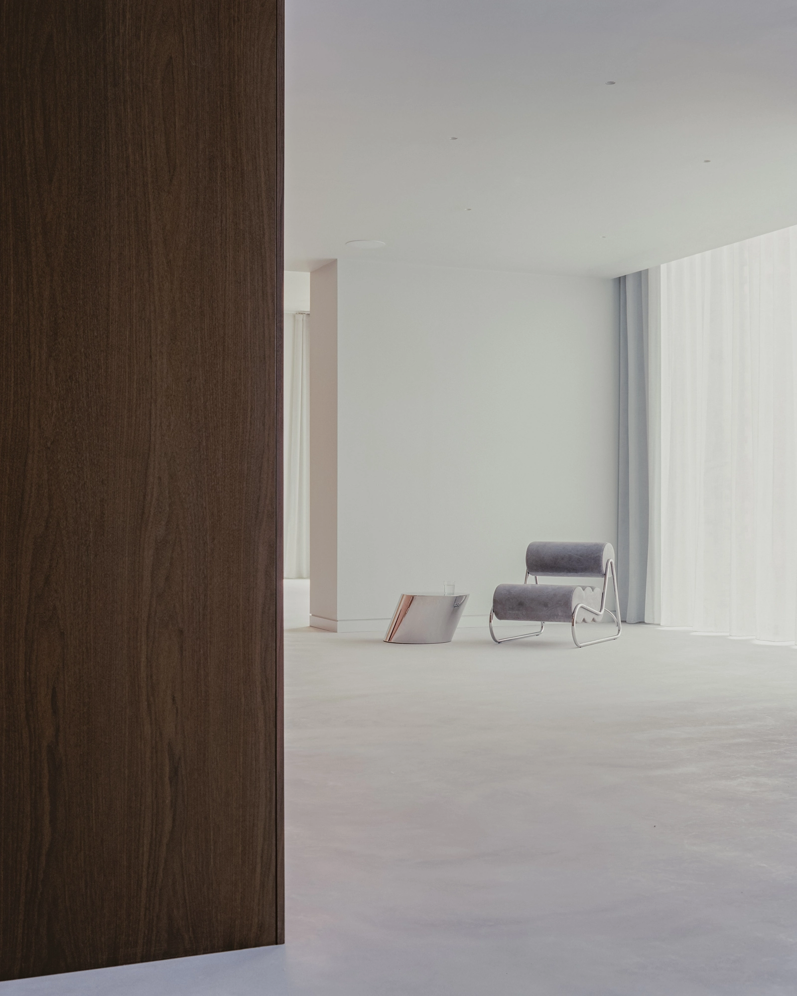 Oval House by Actual Office, photographed by Nick Dearden — A dark walnut panel at left frames the open pale space, where a grey chair and polished chrome side table sit by the curtains.