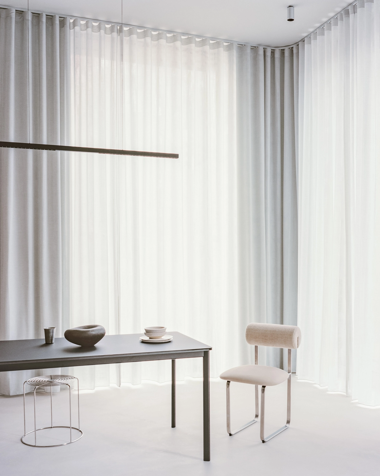 Oval House by Actual Office, photographed by Nick Dearden — Sheer curtains meeting in a corner behind a grey table set with ceramic vessels, a cream chair and a small stool alongside.