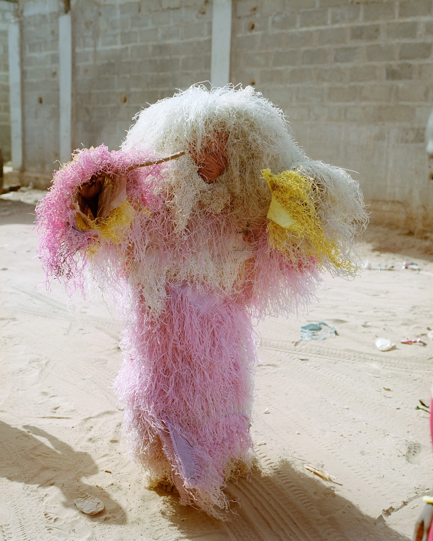 Atlantic Ocean Twilight by Patricia Reyes, photographed by Patricia Reyes — A masked Kankurang figure stands on the sand in front of a block wall, photographed in 2023.