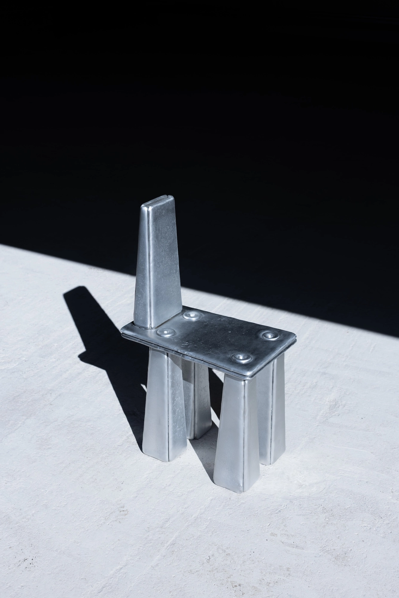 Ingot by Marc Sweeney, photographed by Marc Sweeney — Three-quarter rear view of the chair, the four pour points on the seat plate seen from behind, black ground beyond.