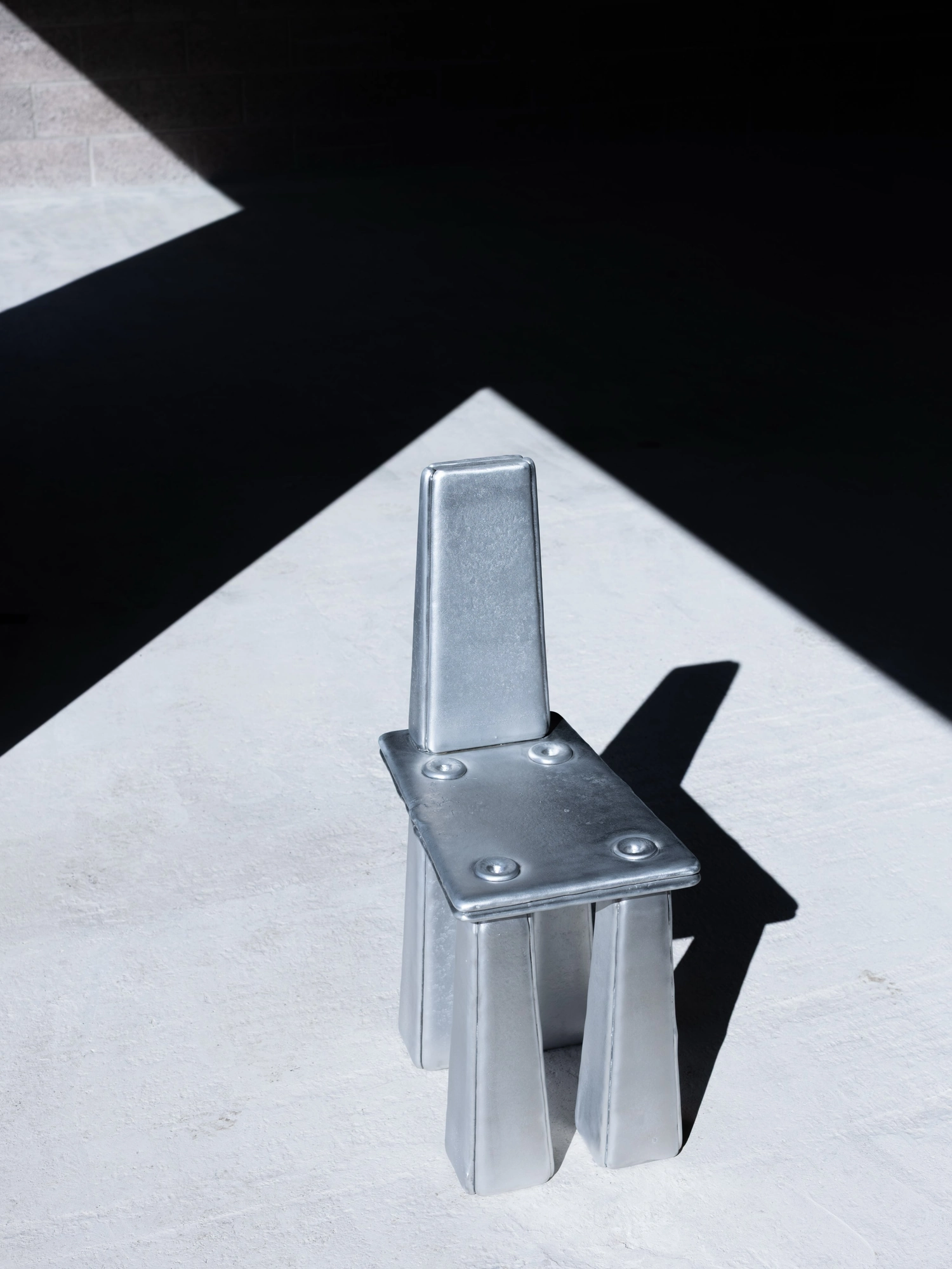 Ingot by Marc Sweeney, photographed by Marc Sweeney — High view of the chair on pale ground cut by an angular black shadow, four pour points across the seat.