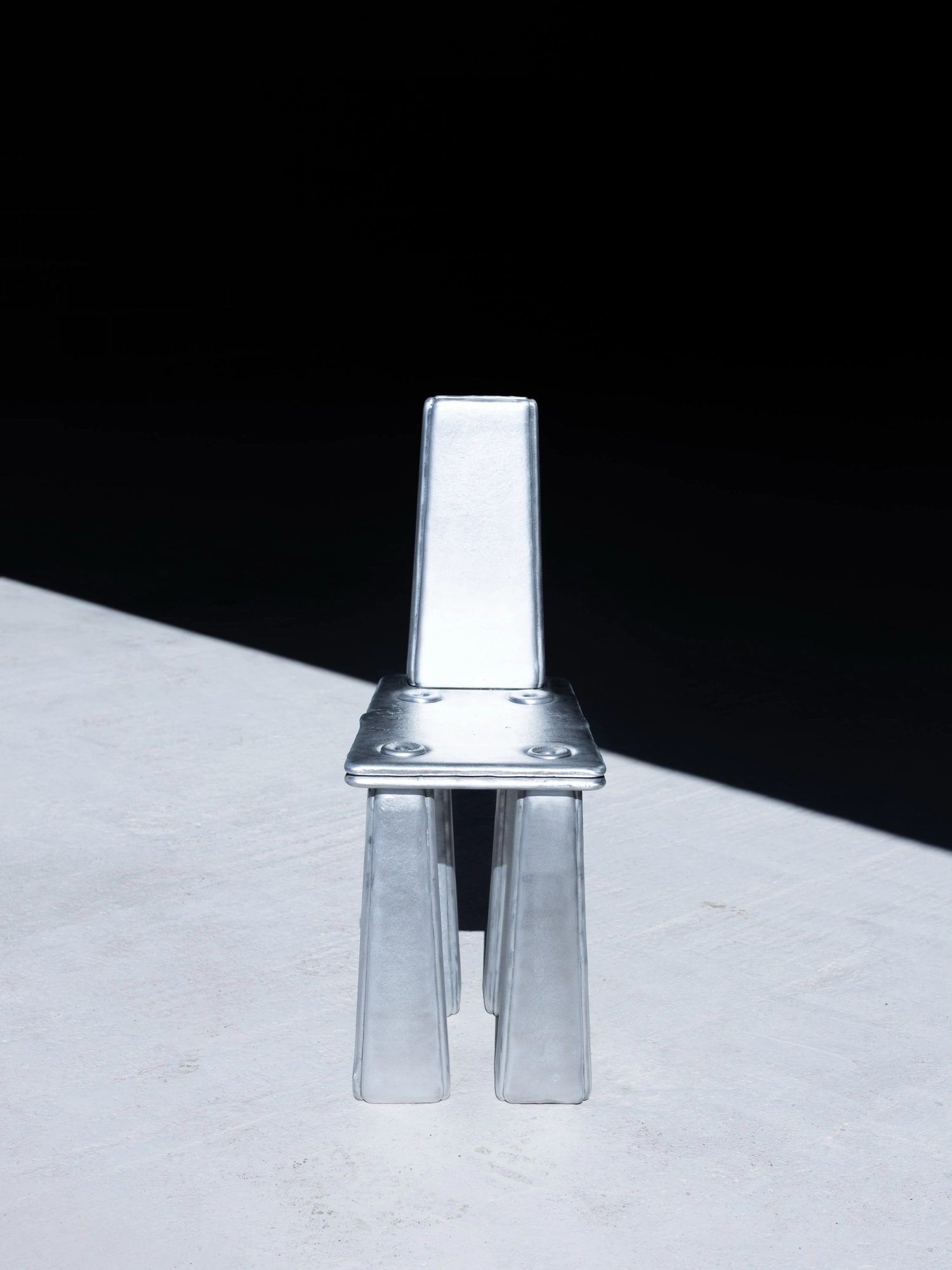 Ingot by Marc Sweeney, photographed by Marc Sweeney — The cast aluminium chair seen from the front, a long diagonal shadow stretching across pale ground, black ground behind.