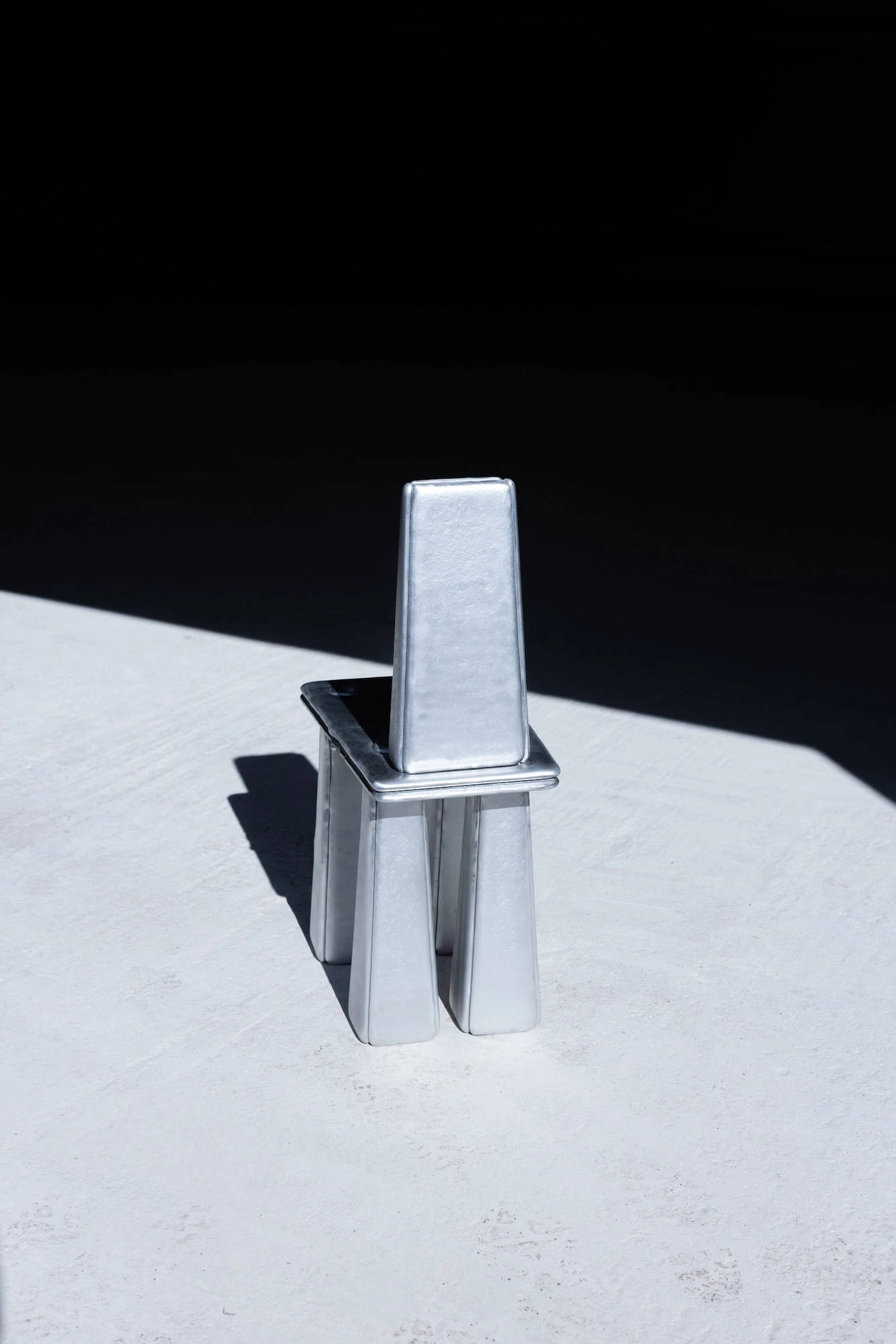 Ingot by Marc Sweeney, photographed by Marc Sweeney — The small cast aluminium chair against a tall black field, legs drawn close, standing in bright sun.
