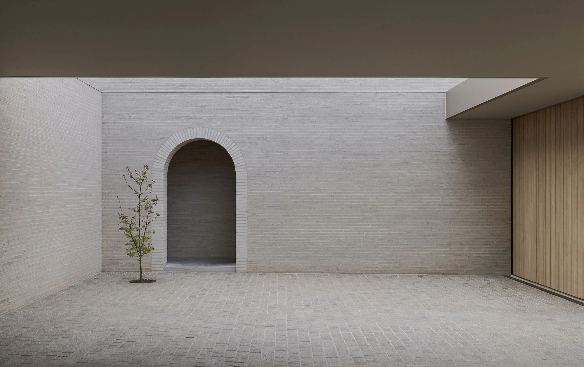 Casa Piva by B.E. Architecture, photographed by Timothy Kaye — A frontal view across the cobbled forecourt to the arched doorway, a young tree standing against the pale brick wall.