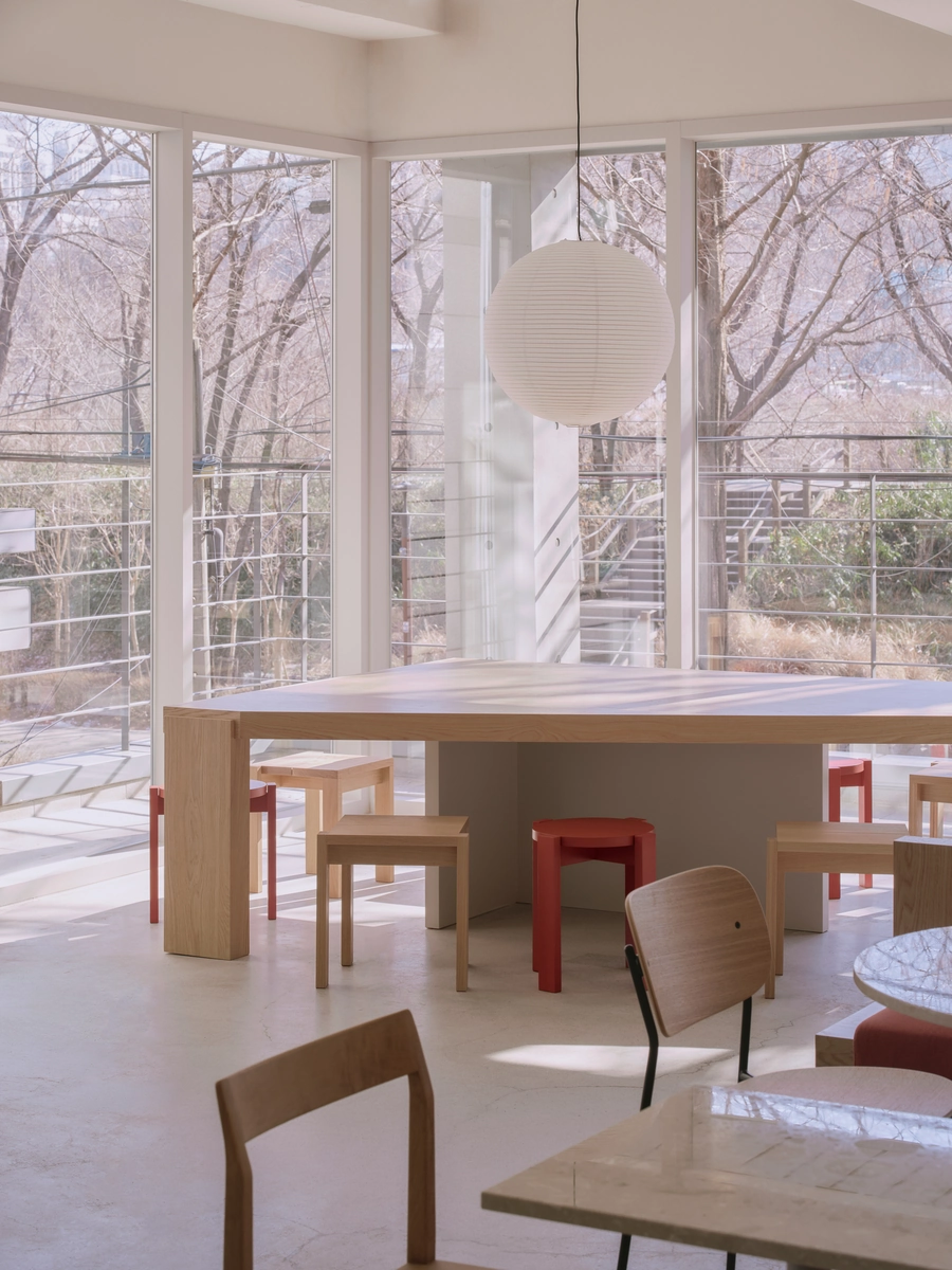 Bread&Walk by StudioFA, photographed by Dongkyu Kim — A communal table meets a corner of full-height windows, stools gathered at its base and round tables alongside, the park's trees beyond the glass.