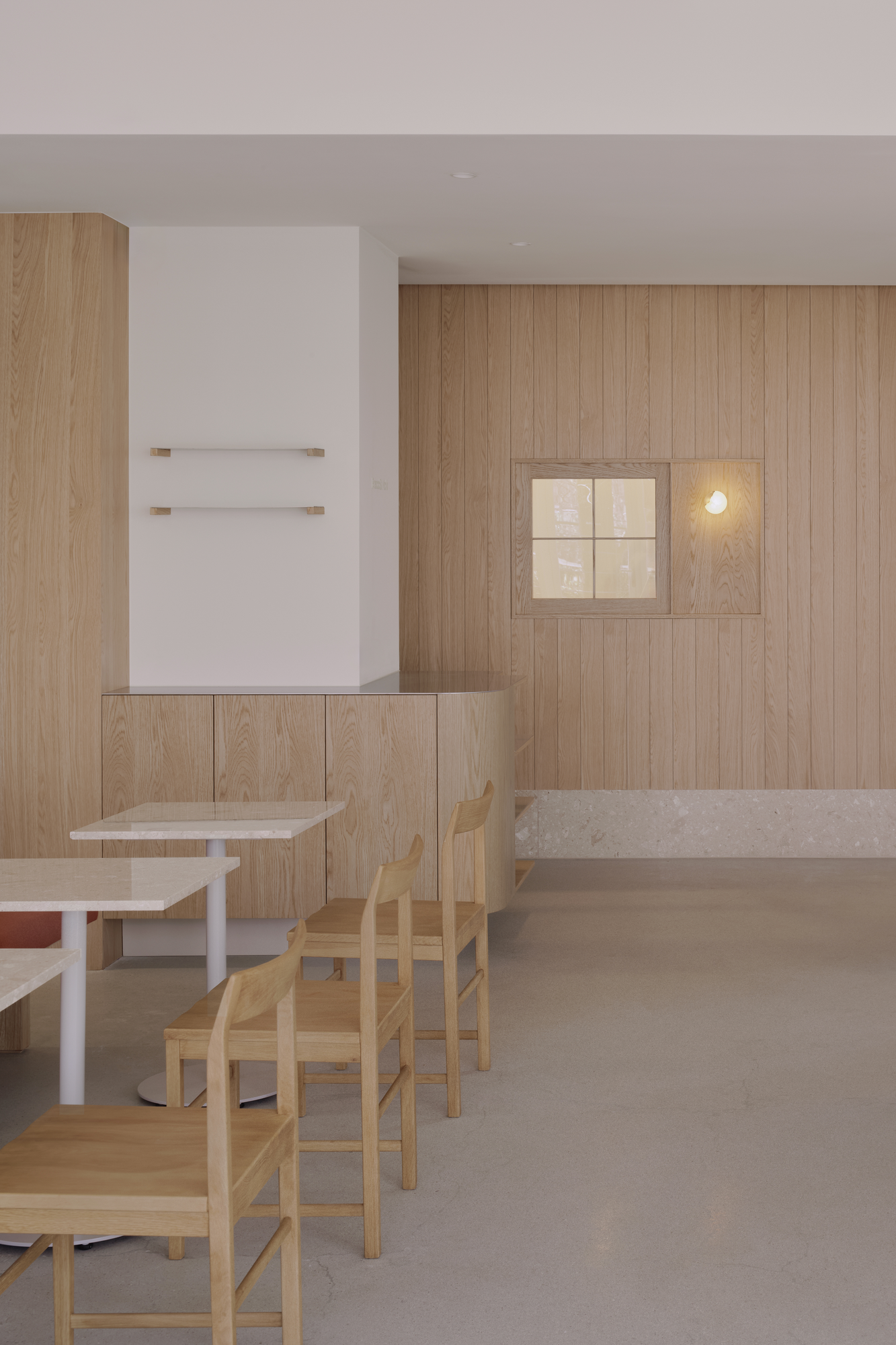 Bread&Walk by StudioFA, photographed by Dongkyu Kim — A square window with a light beside it sits in the wall above wall-mounted shelves and a counter, chairs and tables to the left.