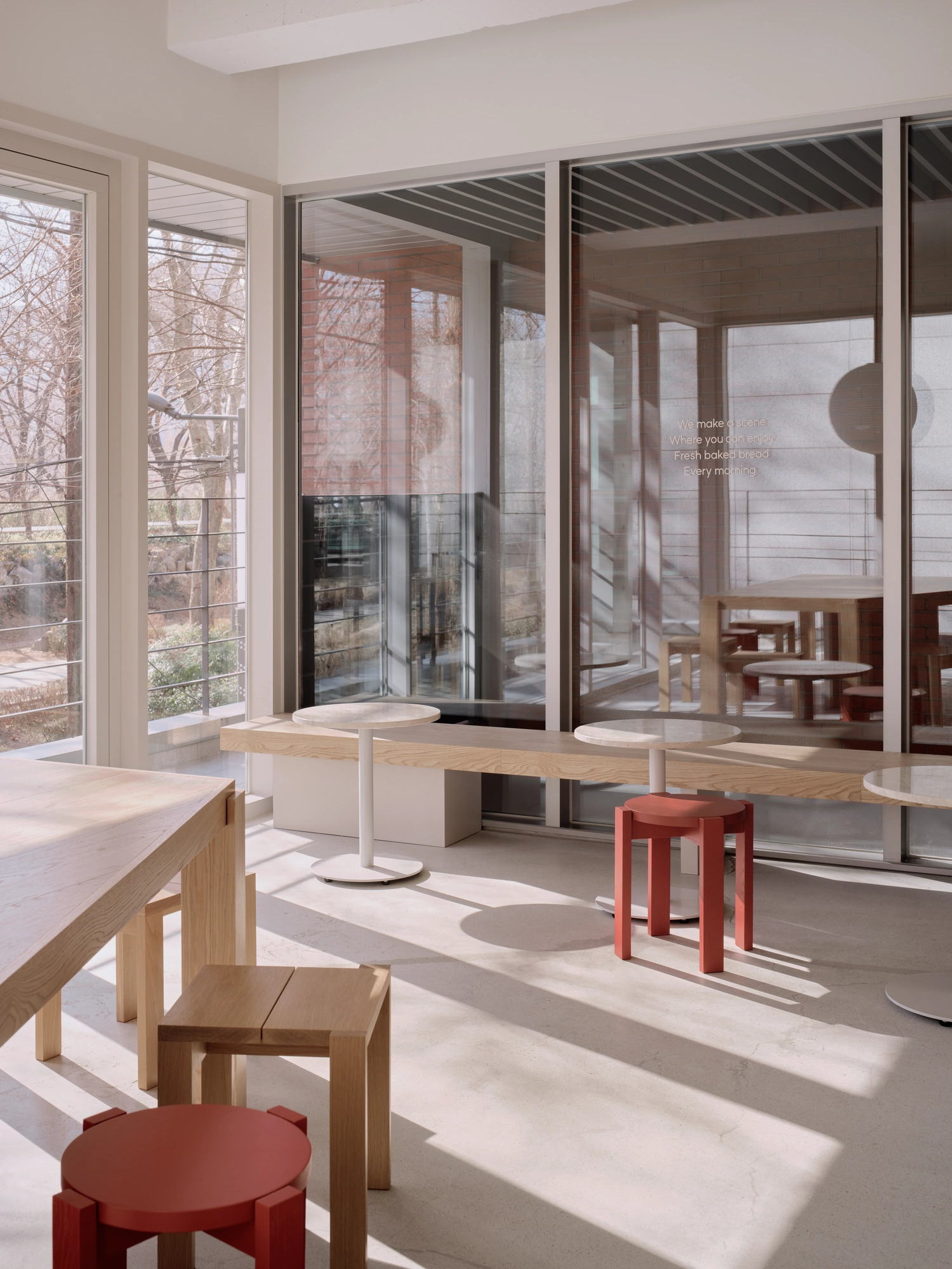 Bread&Walk by StudioFA, photographed by Dongkyu Kim — Tables run along a continuous shelf at the windows with stools beneath, the park and its bare trees seen through the corner glazing.