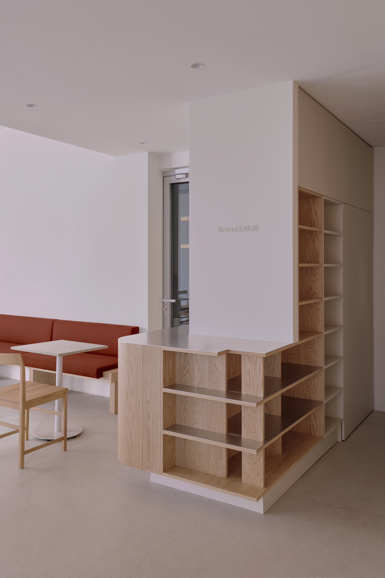 Bread&Walk by StudioFA, photographed by Dongkyu Kim — A bench and a round table sit beside a counter of open shelving, the shop's name set on the wall above.