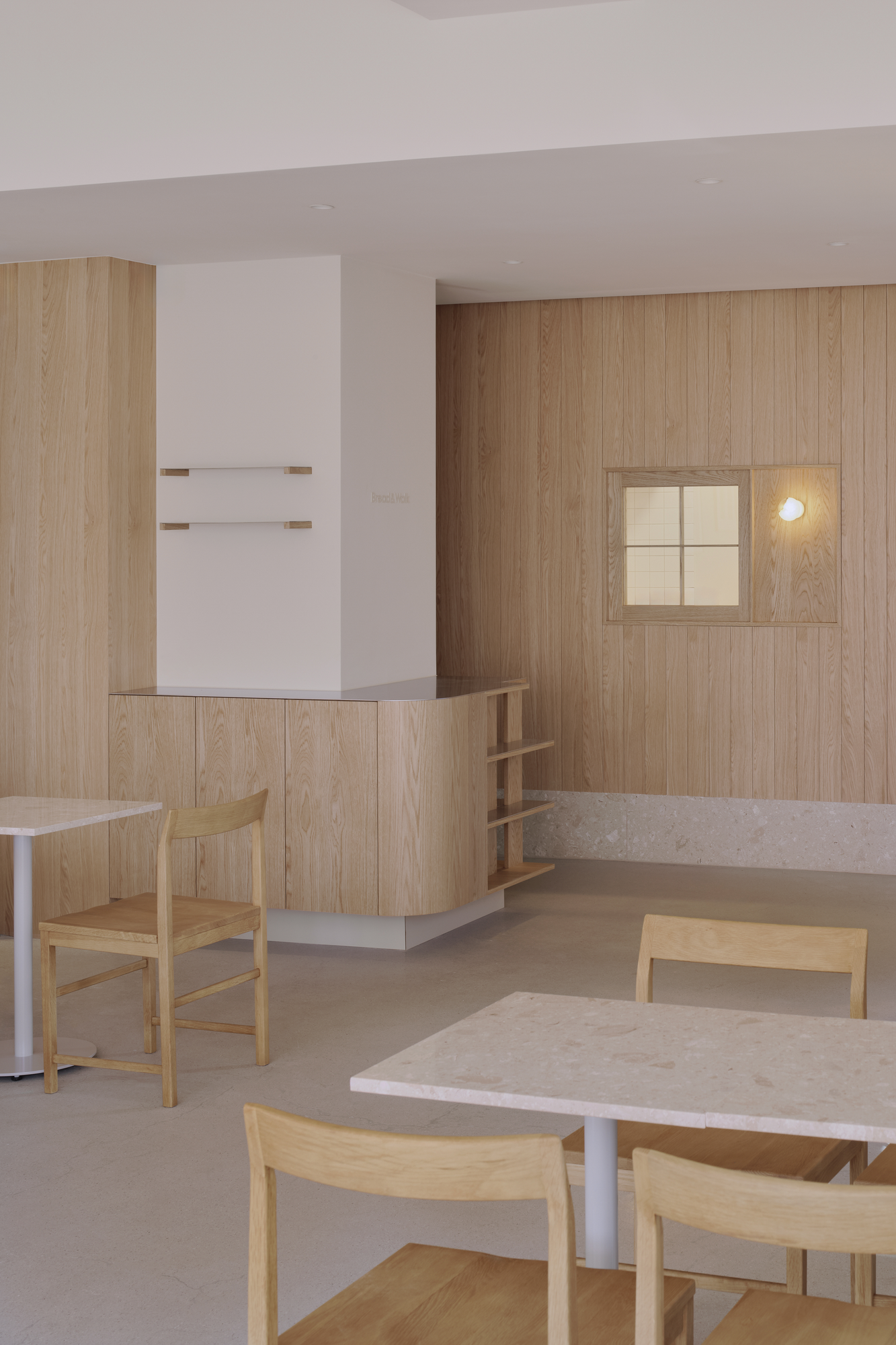 Bread&Walk by StudioFA, photographed by Dongkyu Kim — Thin wall shelves and a counter of open shelving stand under a square window, two tables with chairs across the floor.