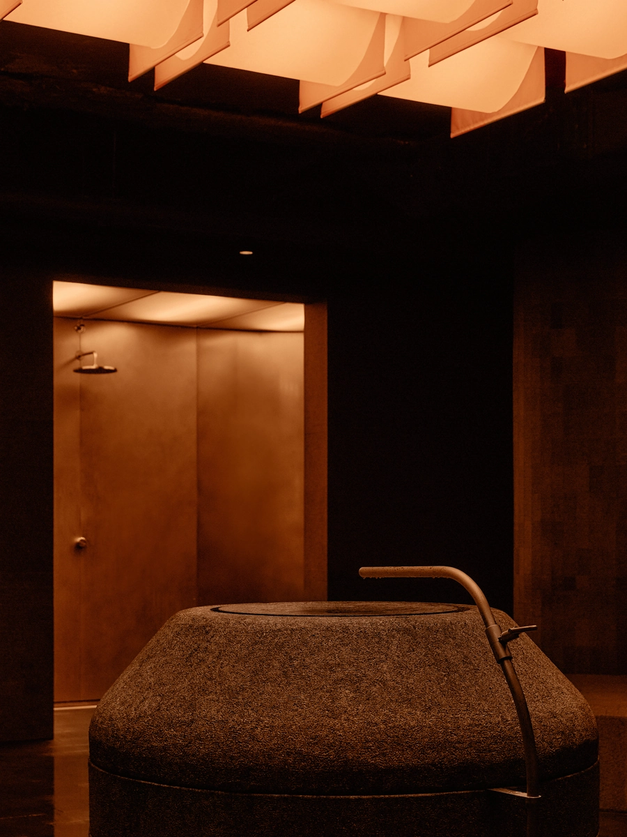 The Altar by MV-LLC — The bush-hammered granite basin where guests draw water, a communal shower lit in the recess behind it.