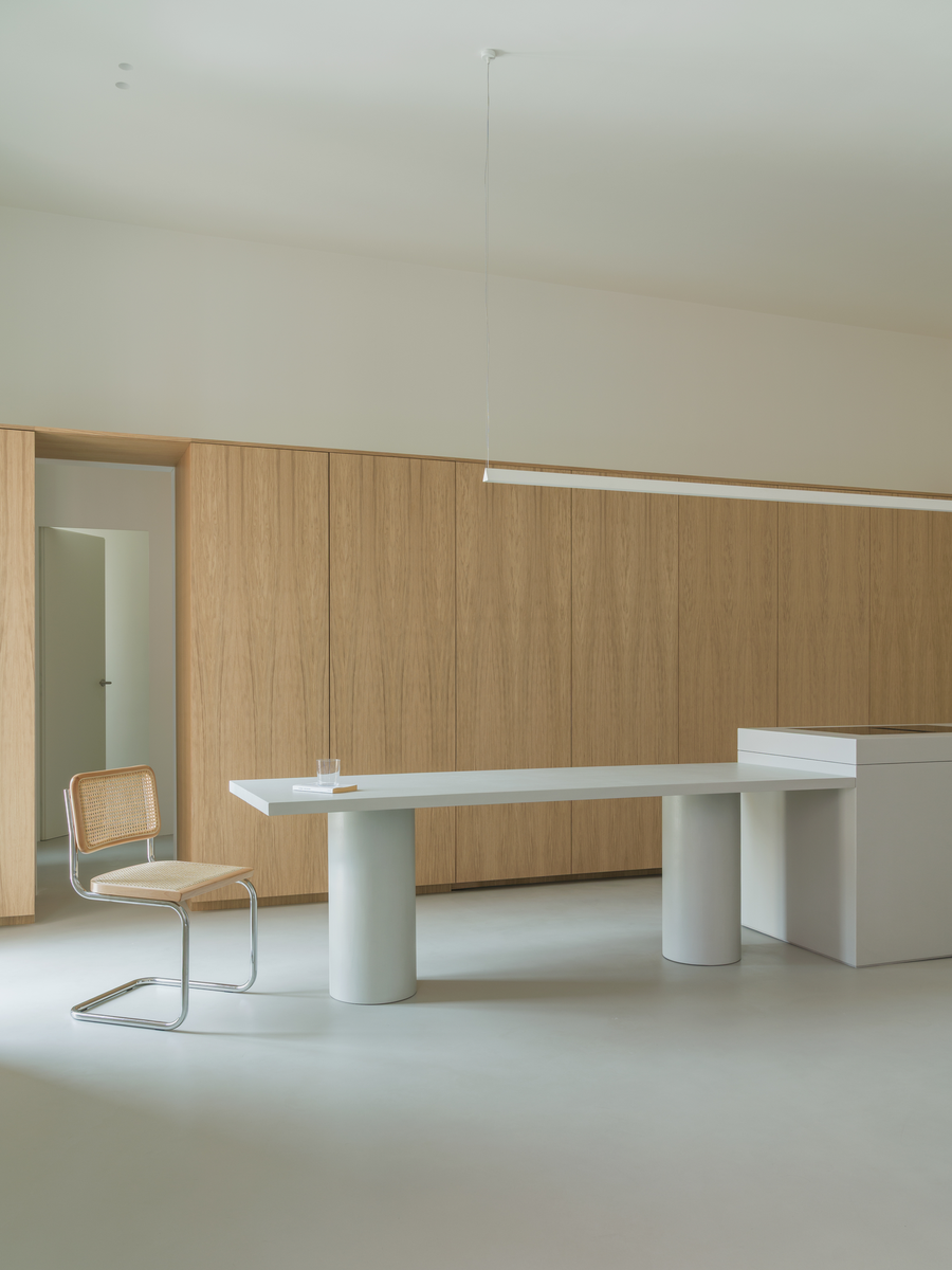House with a White Column by SET Architects, photographed by Daniele Criscenzo — A table on cylindrical legs meets the microcement island in front of a wall of oak doors, with a chair and an opening at the left.