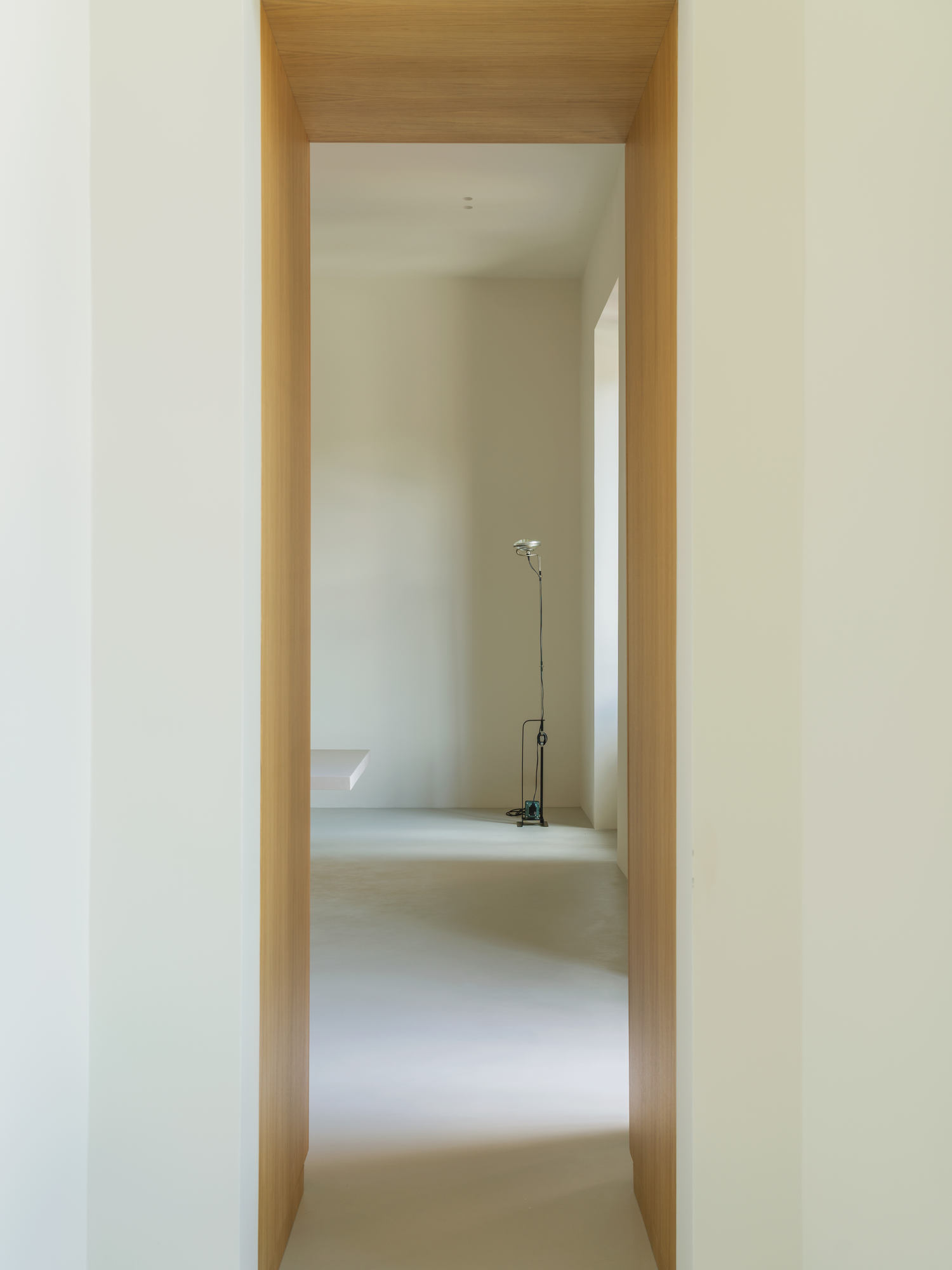 House with a White Column by SET Architects, photographed by Daniele Criscenzo — A doorway lined in oak frames a white room beyond, a floor lamp standing in its far corner on the microcement floor.
