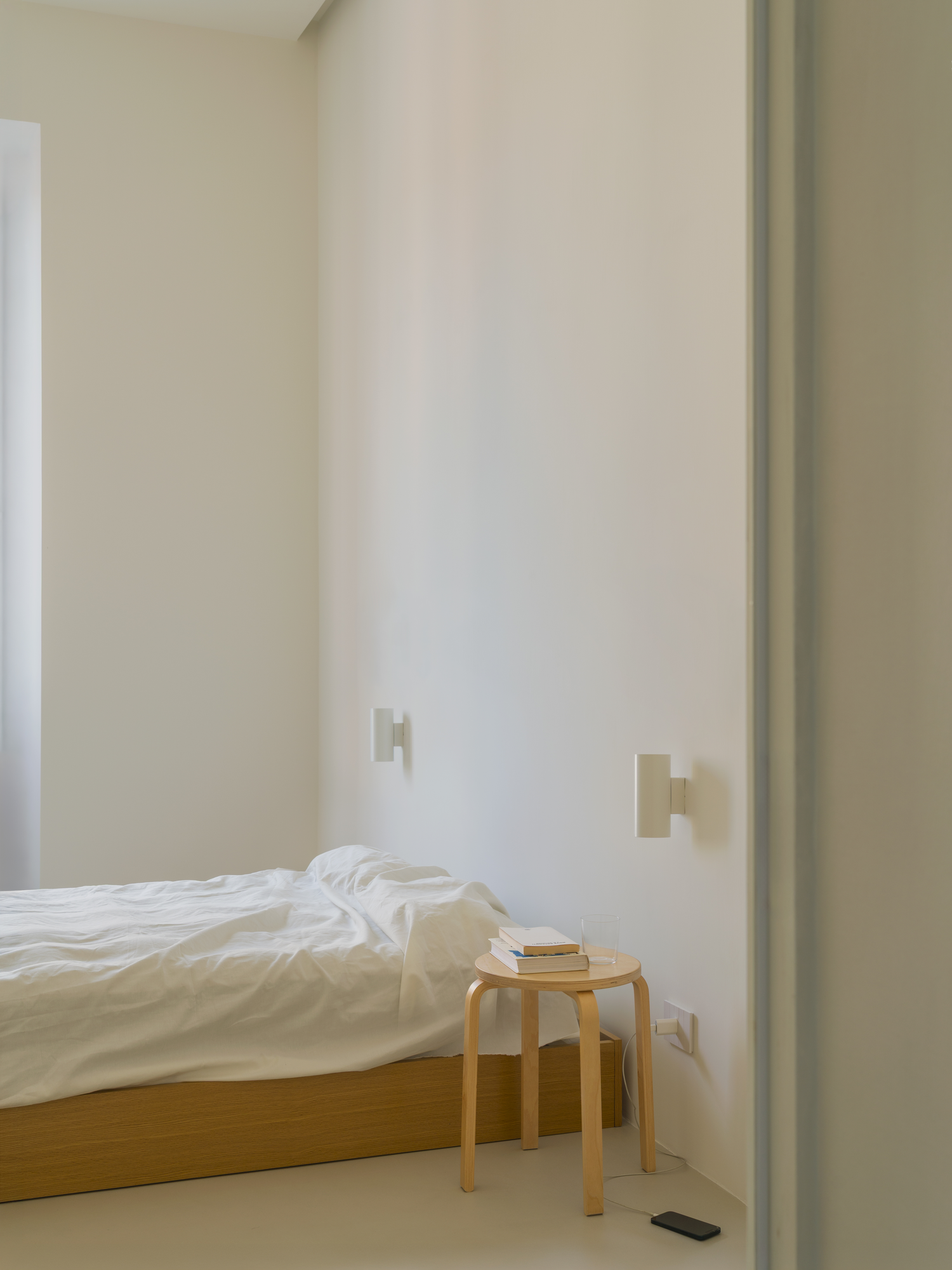 House with a White Column by SET Architects, photographed by Daniele Criscenzo — A bed against white walls, with a stool holding books beside it and wall lights above.