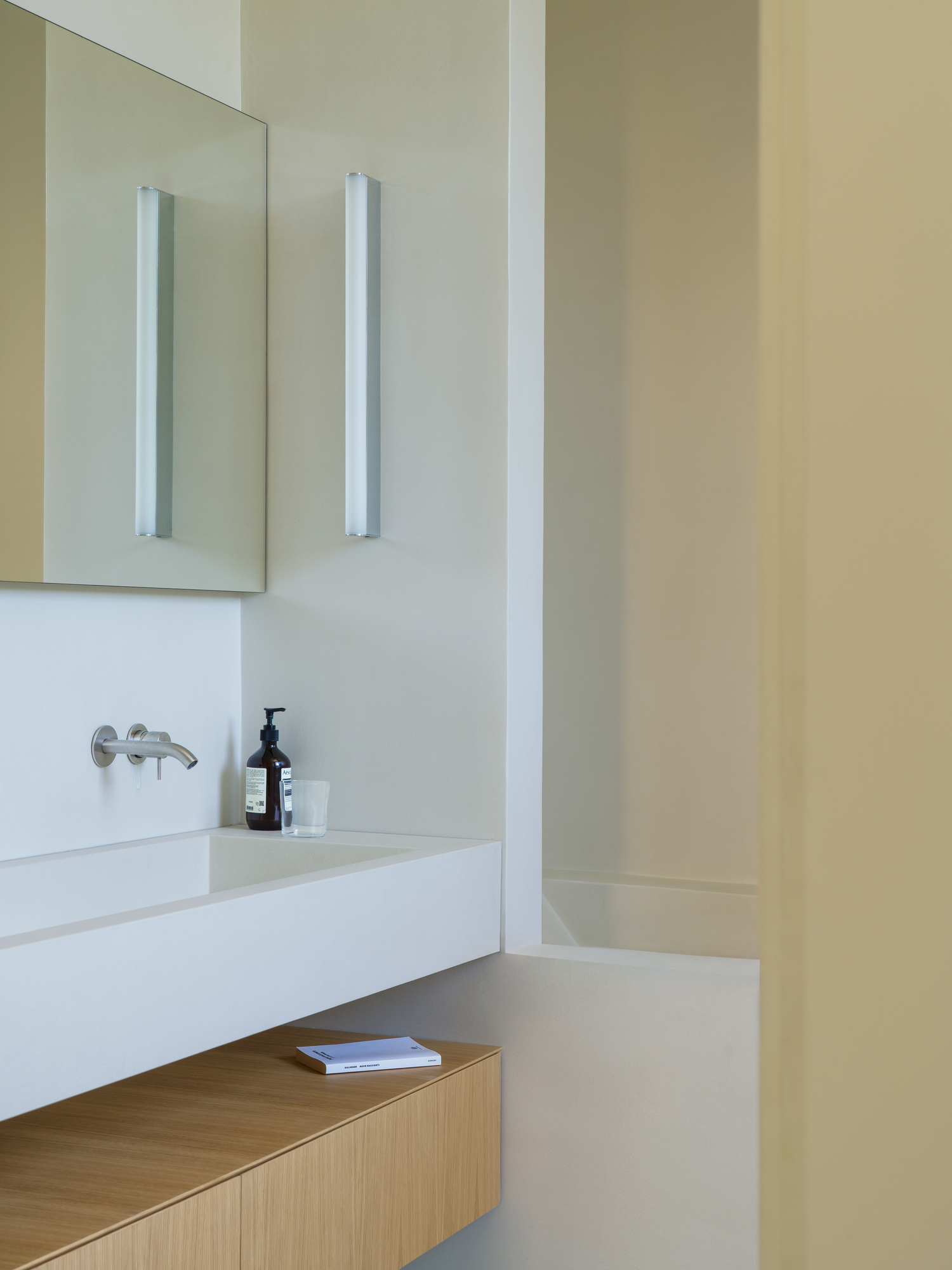 House with a White Column by SET Architects, photographed by Daniele Criscenzo — A bathroom basin under a mirror, with a wall light beside it, a drawer unit below and a recess beyond.
