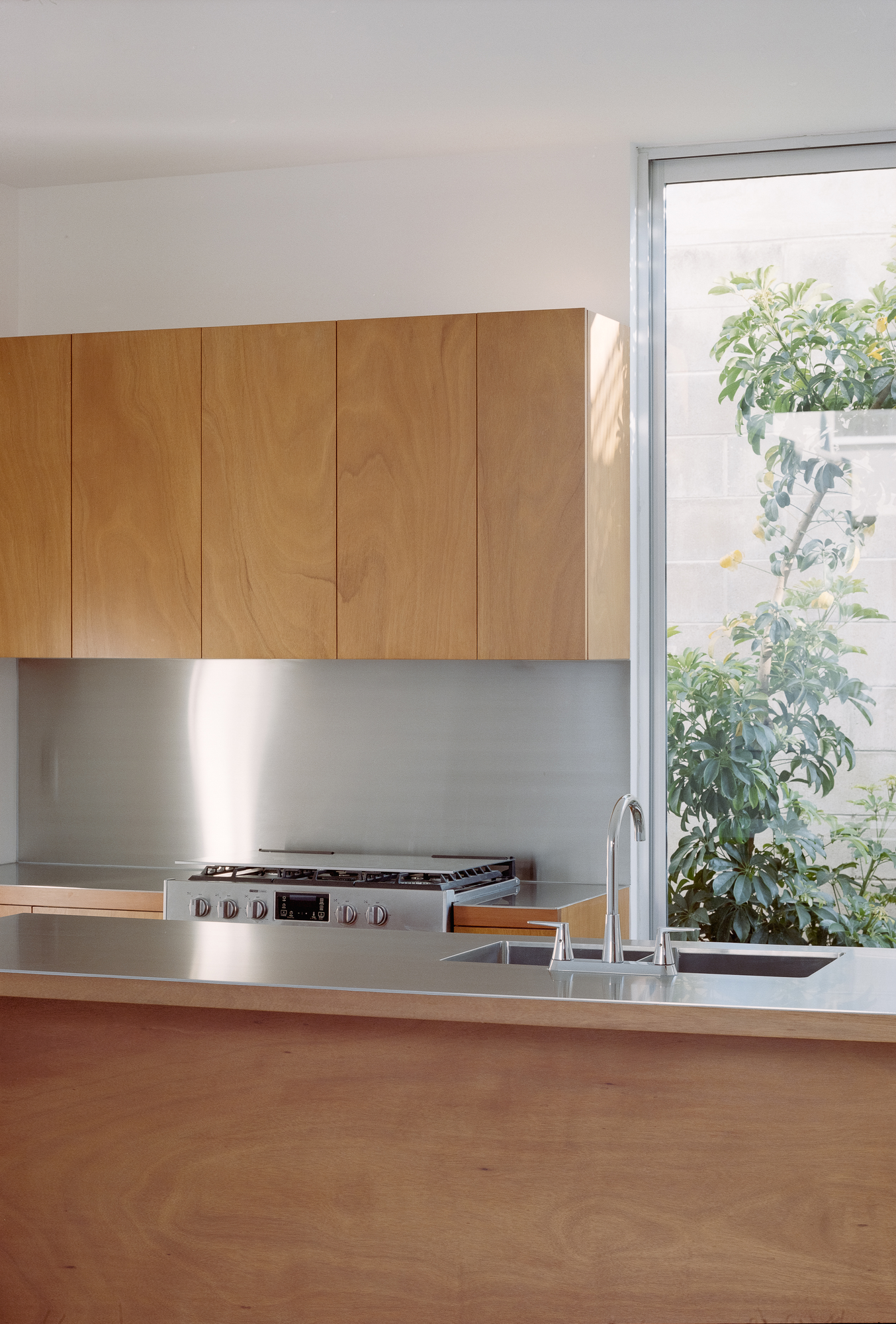 House Within a Cube I by operadora, photographed by Arturo Arrieta — Film photograph of the ground-floor kitchen, a range set into the counter below upper cabinets, a sink with a tap, and a tall window beside a plant.
