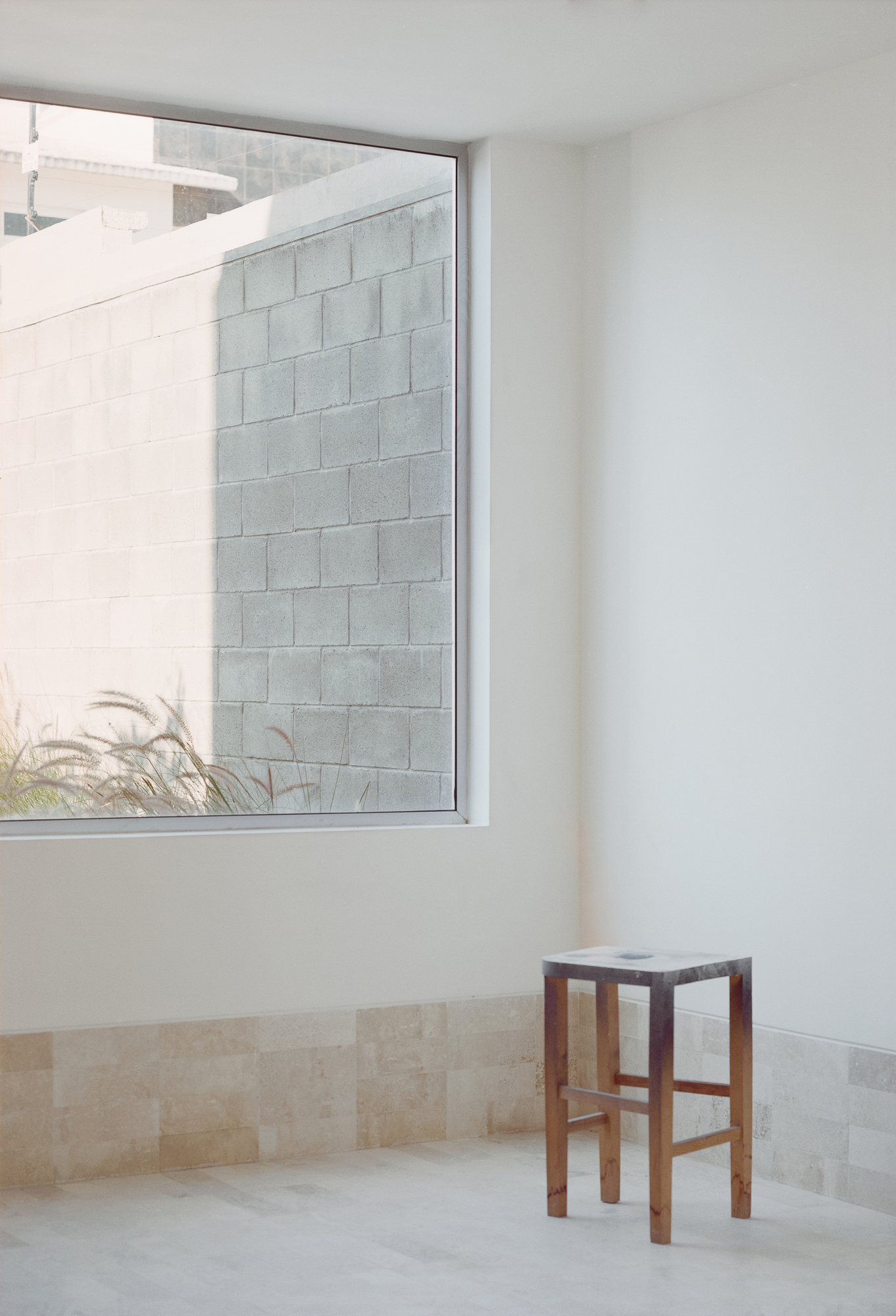 House Within a Cube I by operadora, photographed by Arturo Arrieta — Film photograph of a single stool standing in the corner of a room, a large window at left over grasses and the wall beyond.